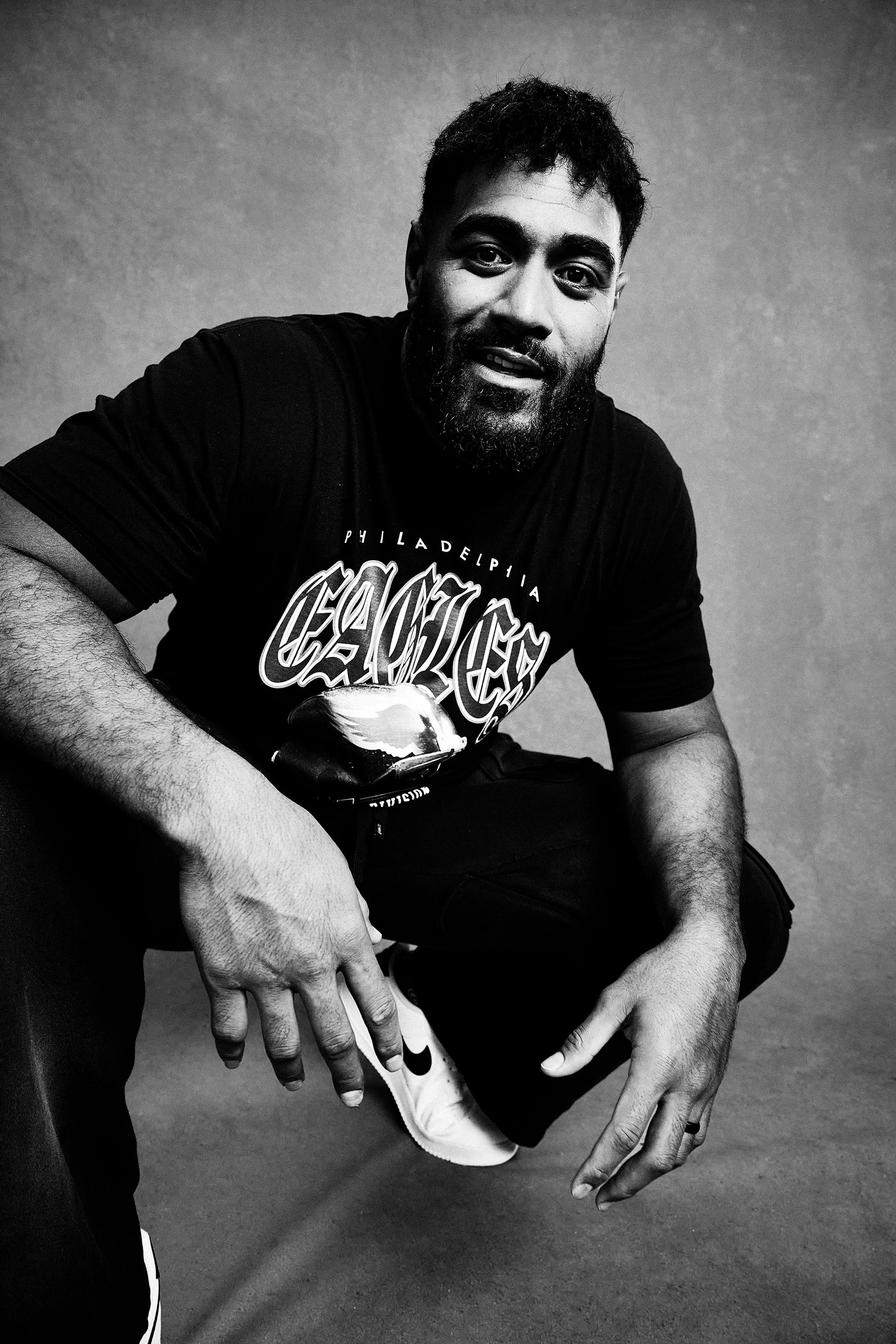 Black and white photo of a bearded man with short hair, wearing a T-shirt with 'Philadelphia Eagles' logo and white sneakers, sitting or crouching against a plain background.