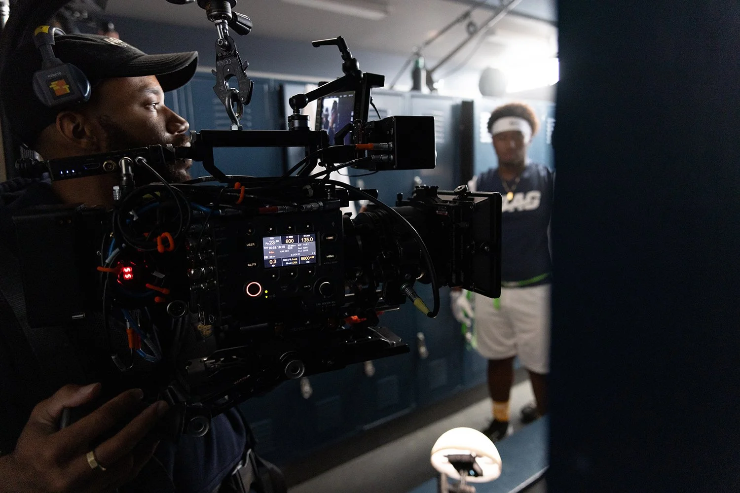 A person operating a professional film camera on set with a tennis player in the background.