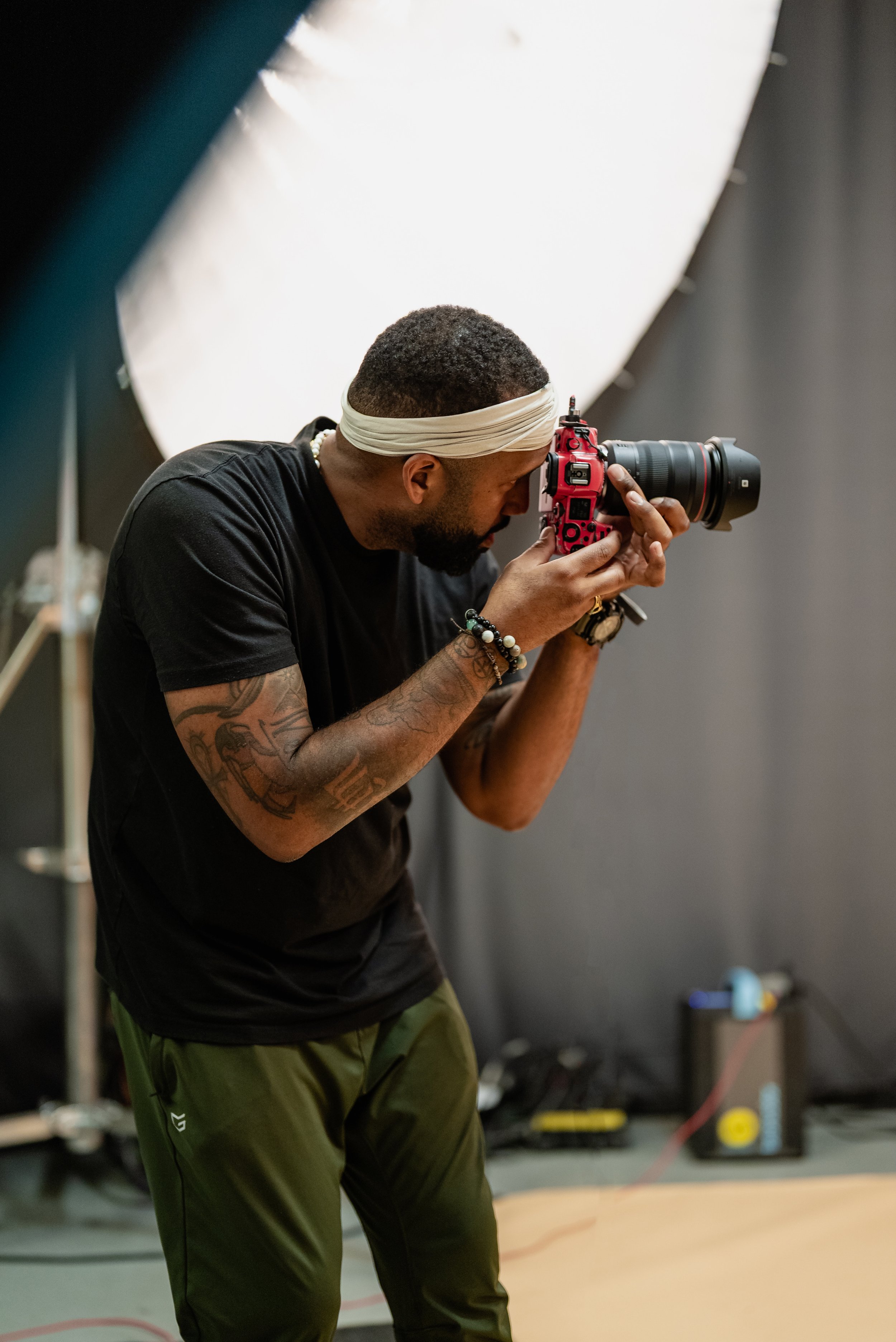 A man with tattoos, a white headband, and a black shirt is taking photos with a red and black camera in a studio setting.
