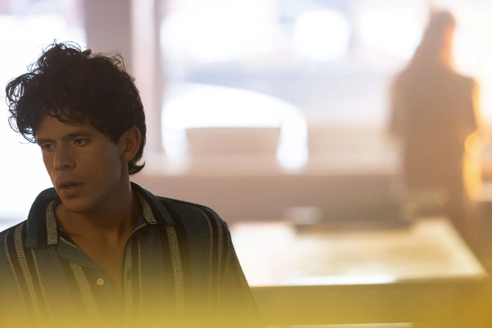 A man with dark, curly hair and light skin looking to the side in a room with bright natural light, wearing a dark shirt with light stripes.