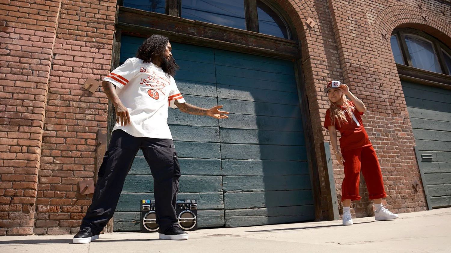 A man and a woman dancing outdoors in front of a brick building with blue garage doors. The man has curly hair and is wearing a San Francisco 49ers football jersey, black pants, and white sneakers, and is gesturing with his right hand. The woman is w