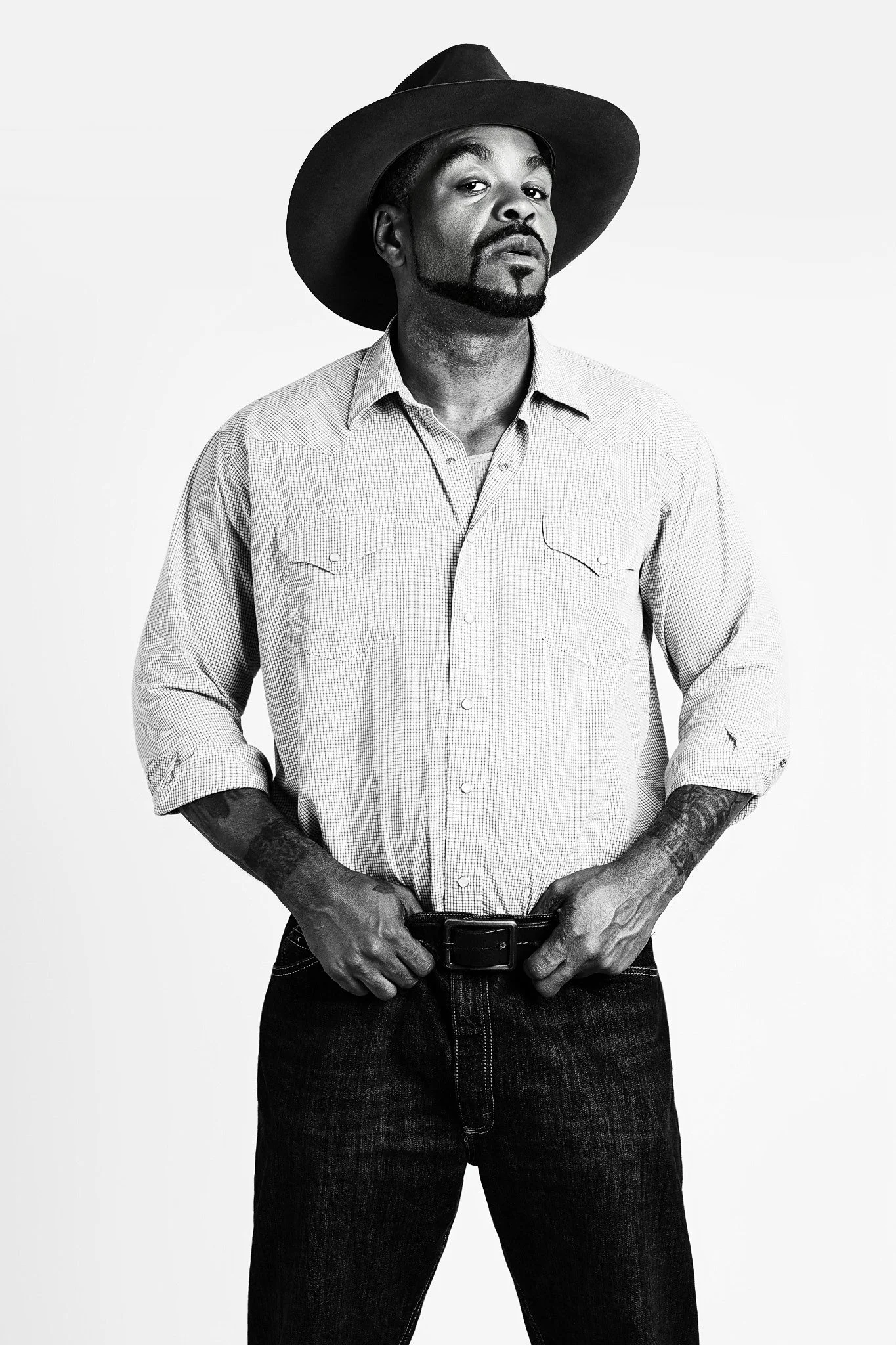A black-and-white portrait of a man wearing a wide-brimmed hat, a checkered button-up shirt with rolled sleeves, and dark jeans, standing with hands on his belt.