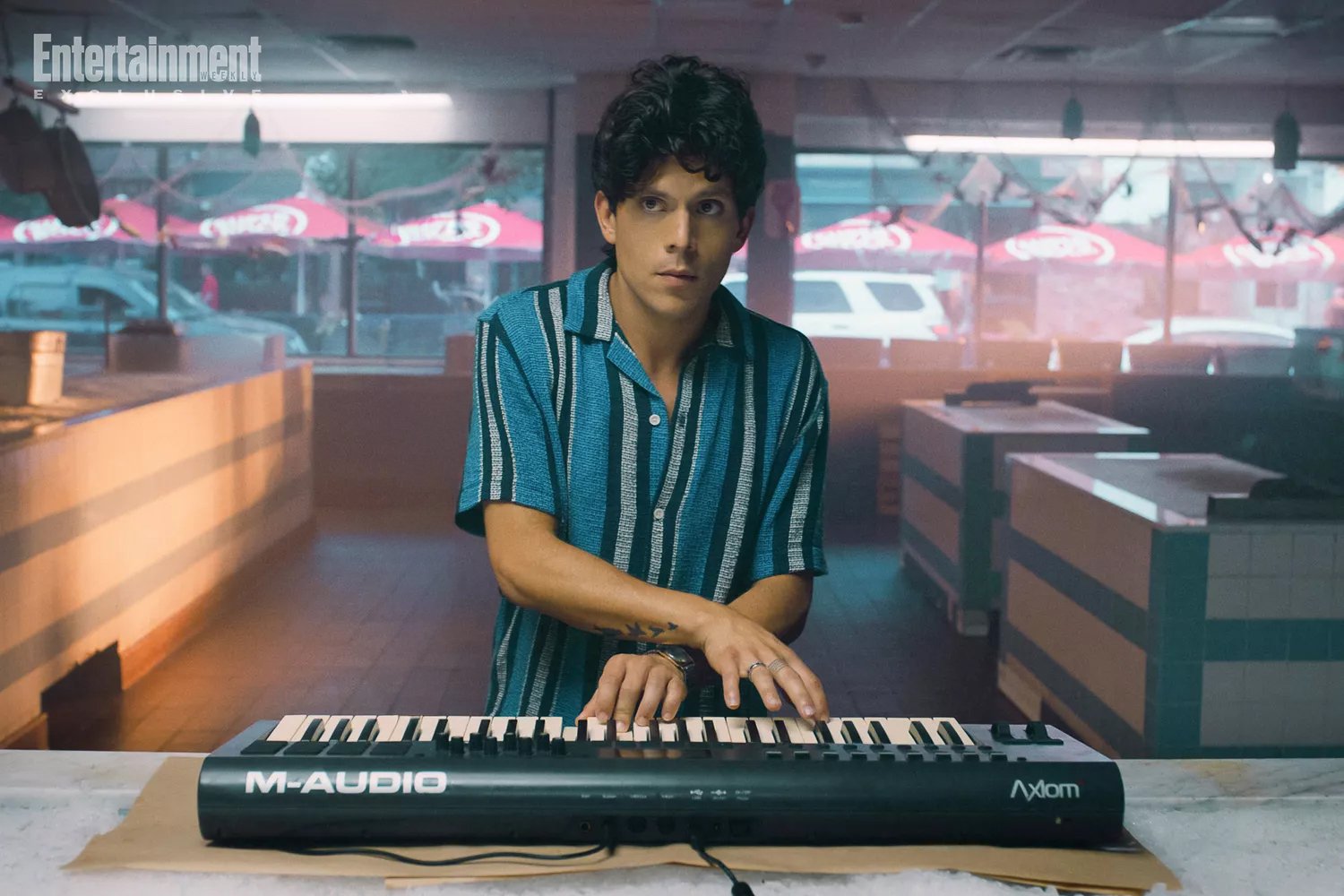 A man with dark curly hair in a striped shirt playing a keyboard in a dimly lit restaurant or cafe, with outdoor seating visible through large windows in the background.