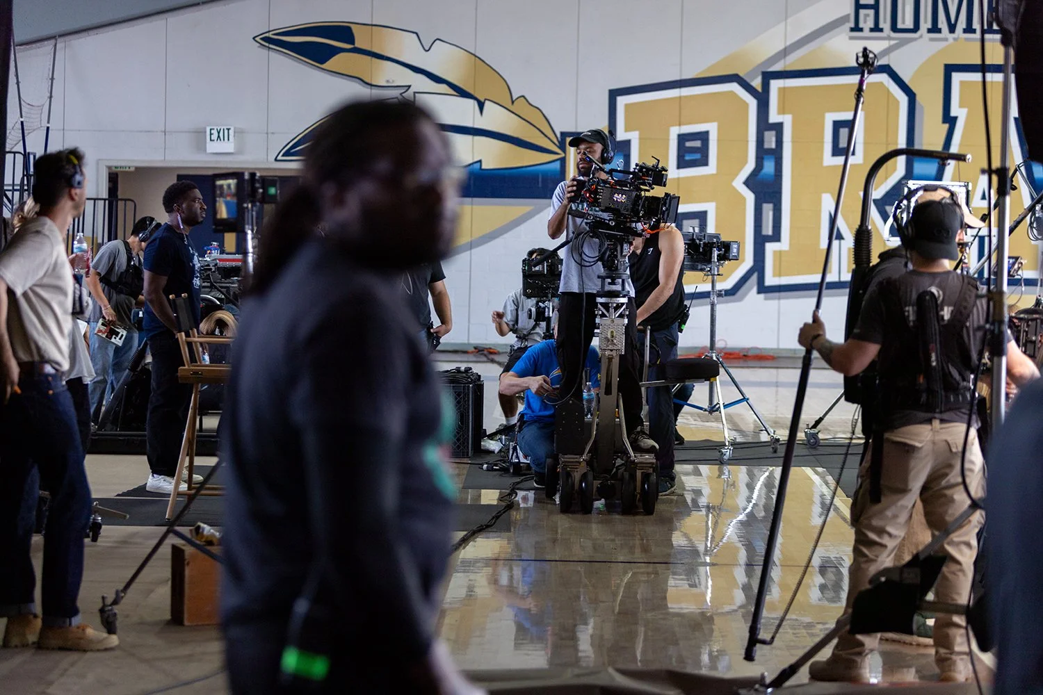 Film crew setting up camera equipment in a gymnasium with a large wall graphic featuring a pen and large letters in the background.