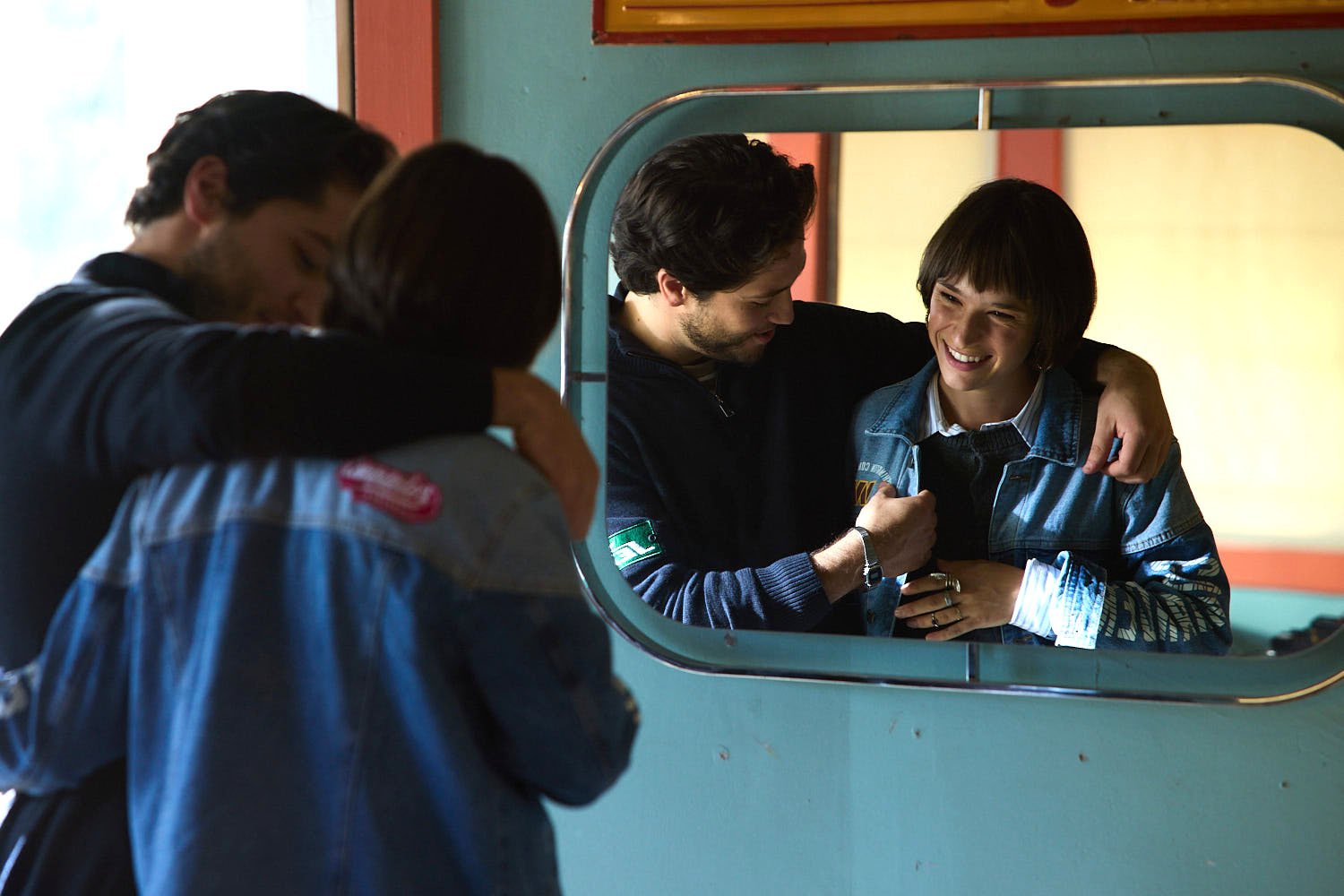 Four people are sitting together in a room, near a mirror. They appear to be happy and sharing a moment of closeness, with two of them having their arms around each other. The room has teal walls and a window in the background.
