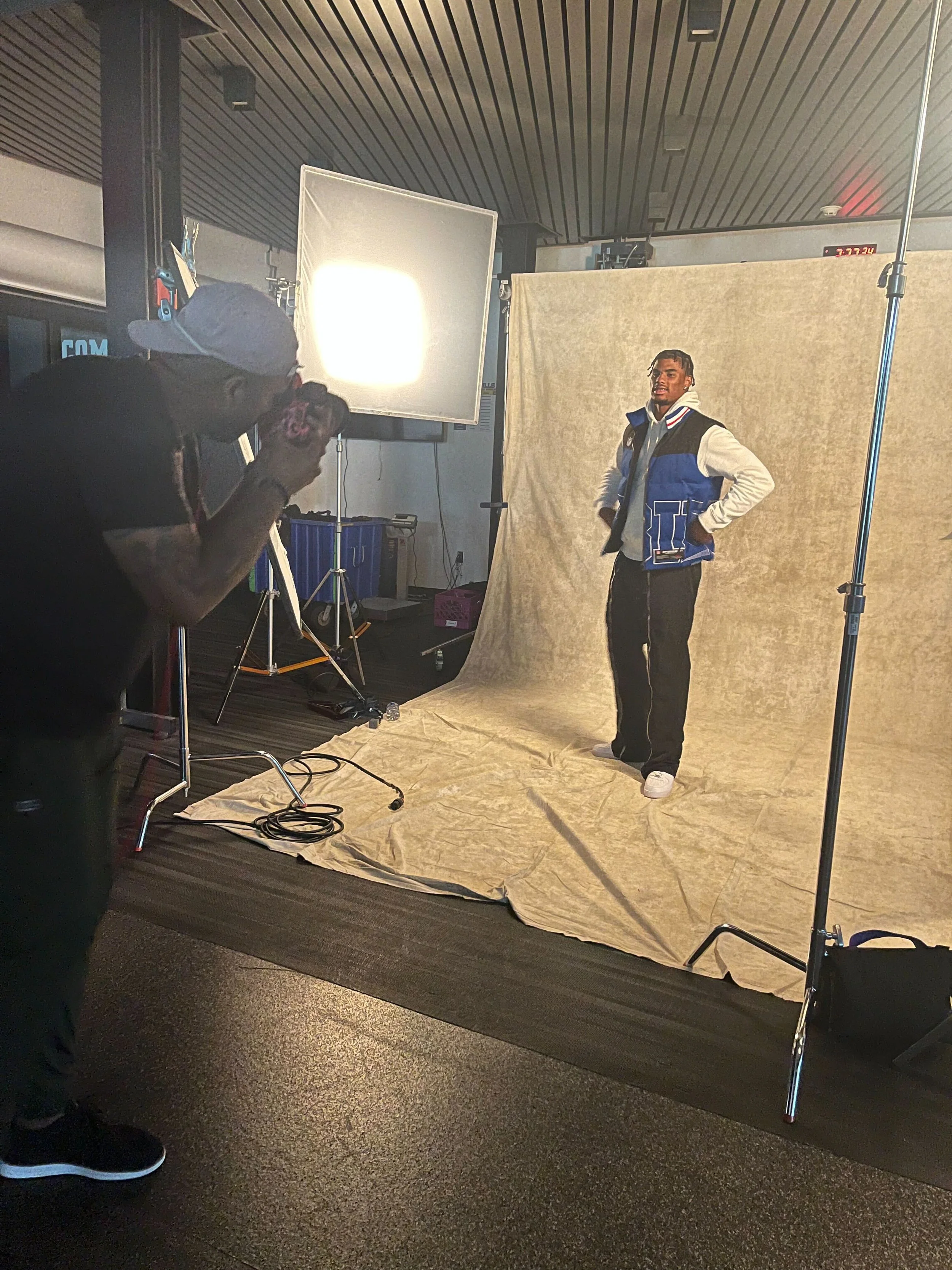 A photo of a photo shoot showing a man posing in front of a beige backdrop with studio lights and equipment. A photographer is taking pictures of him.