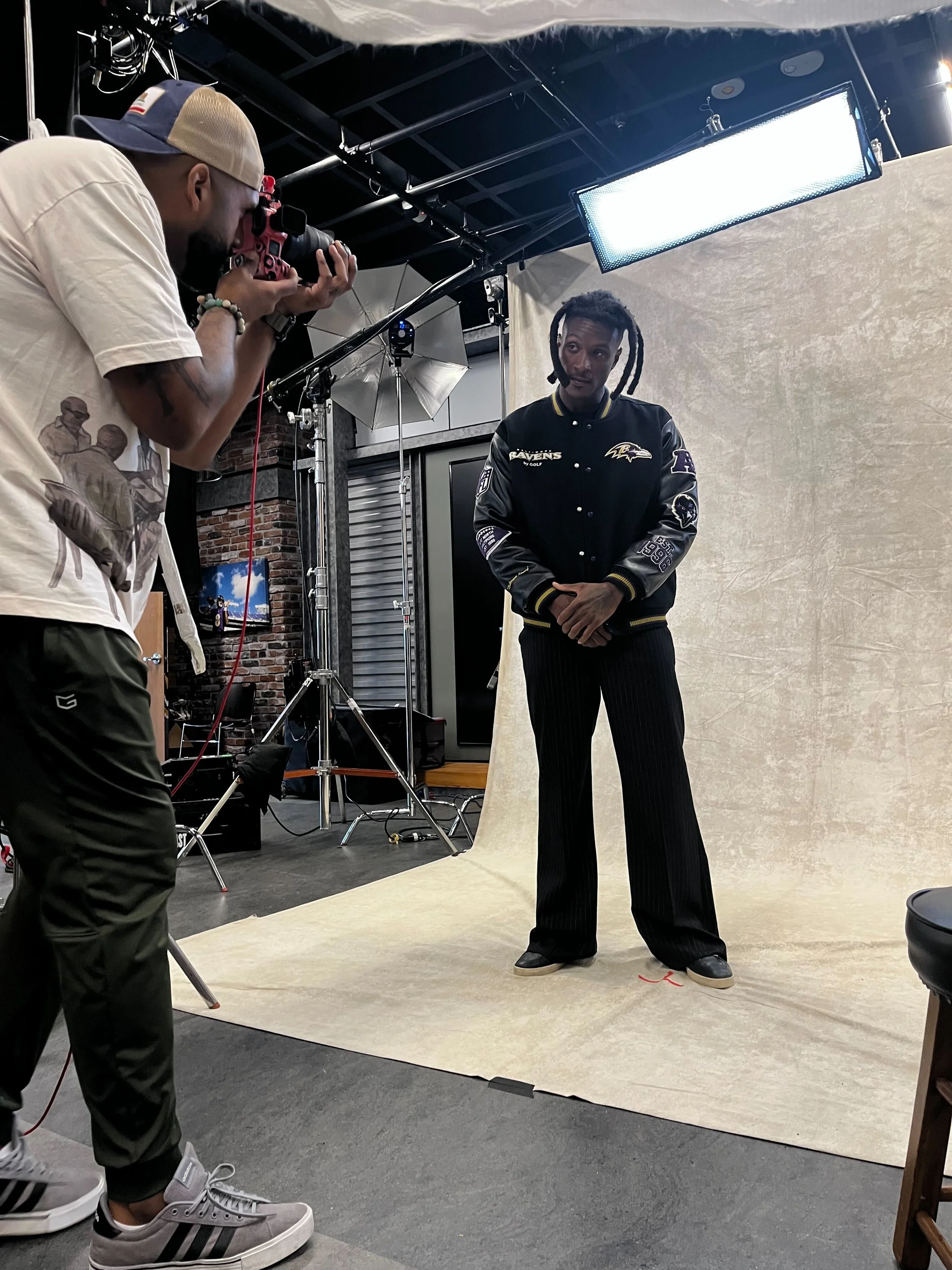 A man with styled dreadlocks dressed in a black Baltimore Ravens jacket and black wide-leg pants stands against a beige backdrop on a photography set, while a photographer with a beige cap and casual clothing takes pictures of him.