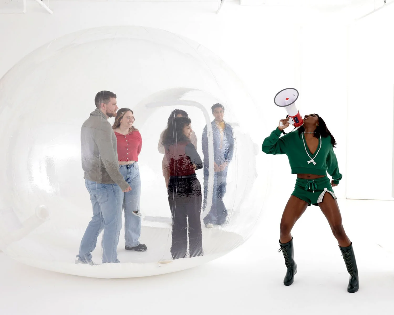 Person in green athletic outfit with boots using a megaphone in front of a clear bubble with people inside.