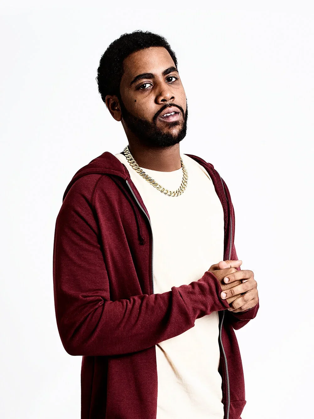 A young man with dark curly hair and a beard, wearing a white t-shirt, a gold chain necklace, and a maroon hoodie, standing against a plain white background.