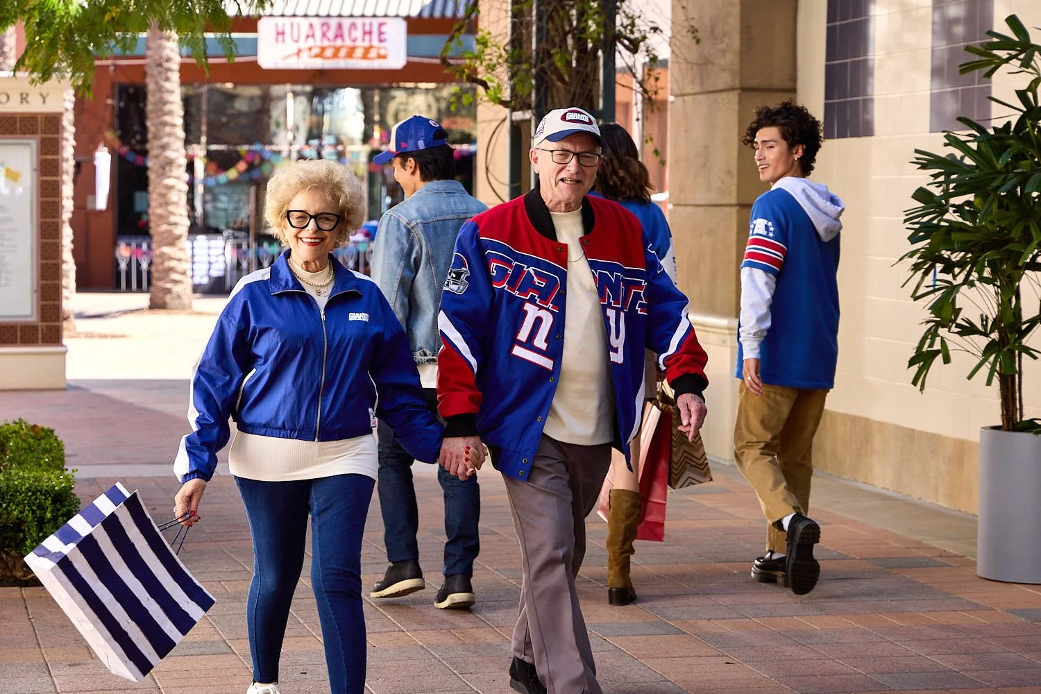 A group of people walking in a shopping mall, dressed in New York Giants sports gear. An elderly woman with glasses and a blue jacket is holding a striped shopping bag, smiling with a man in a Giants jacket. Other individuals are seen in the backgrou