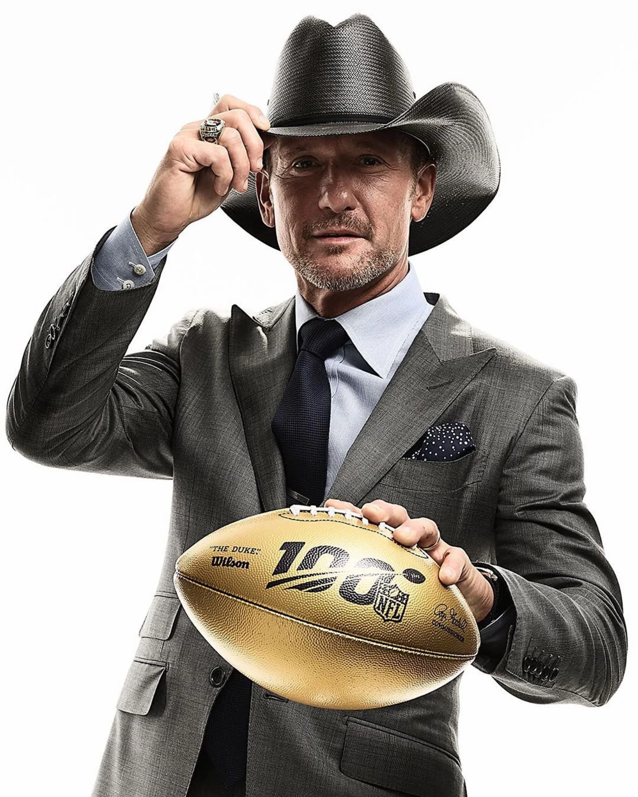 A man in a gray business suit with a blue dress shirt, tie, and pocket square is holding an American football and tipping a black cowboy hat. The football has NFL branding and the number 100.
