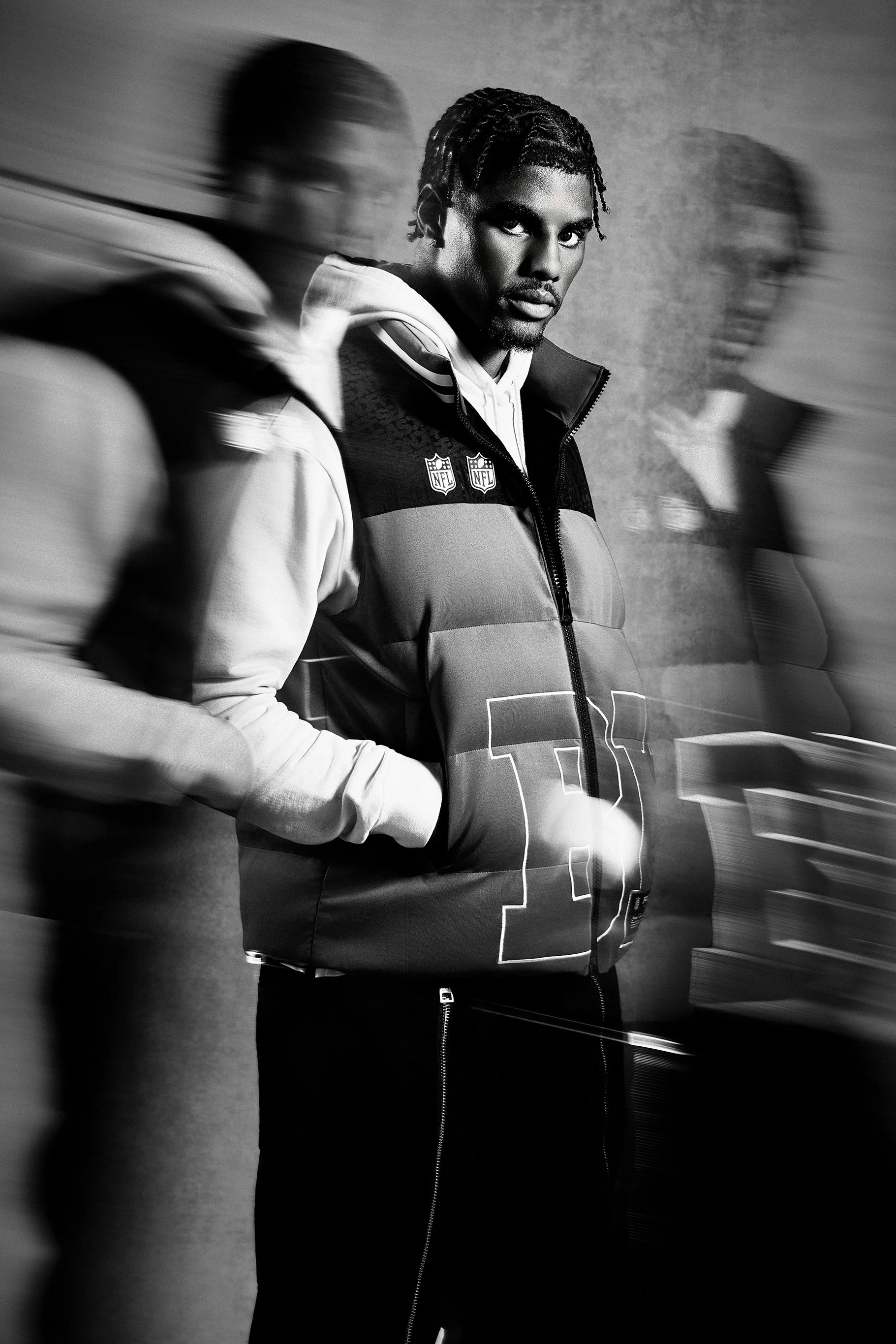 A young man with dark skin, styled dreadlocks, wearing a sports jacket with NFL logos, standing against a textured wall, with two blurred figures of men on either side.