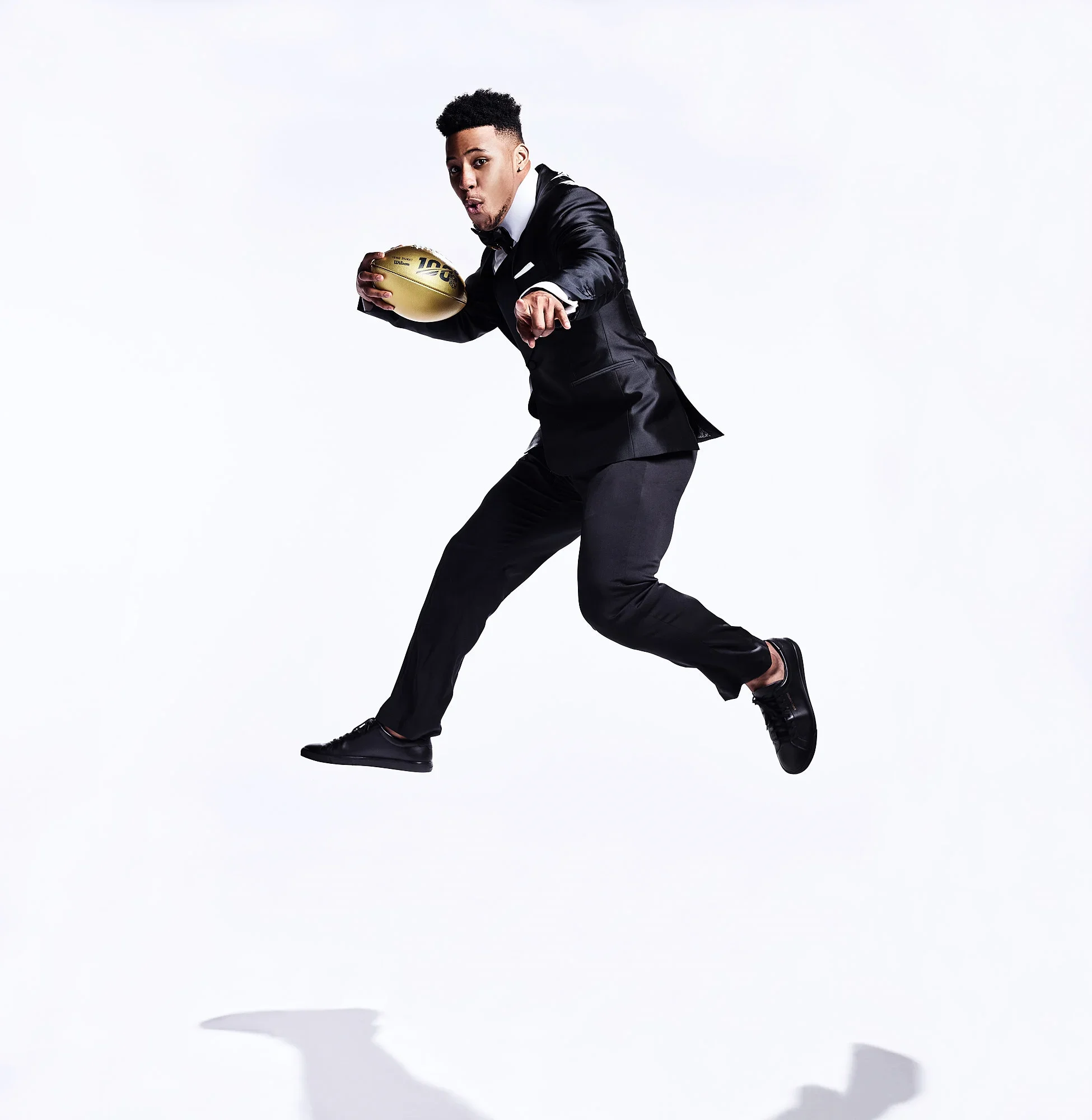 Man in a tuxedo holding an American football, in mid-air as if running or jumping against a white background.