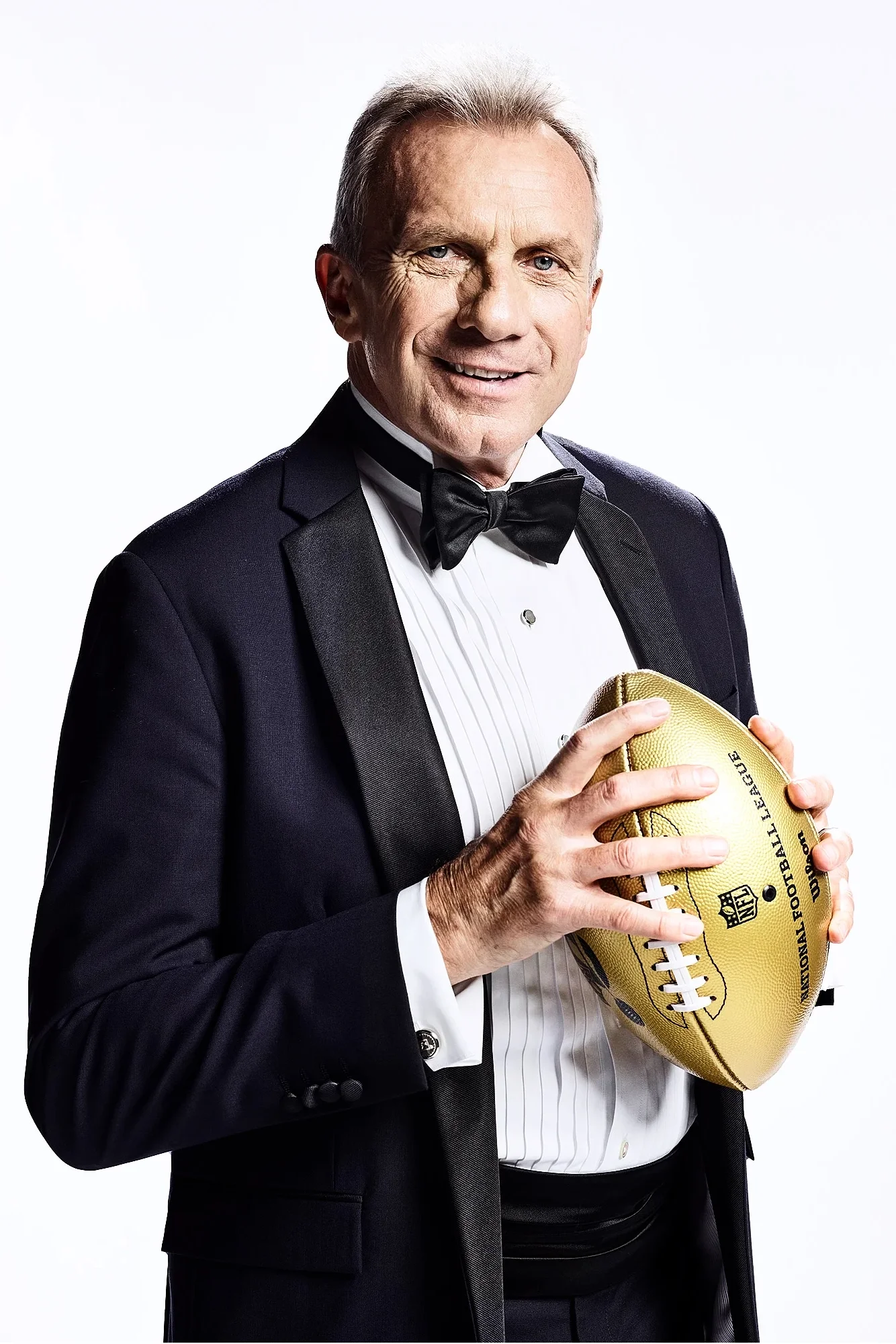 A man in a tuxedo holding a gold football, smiling, with a white background.