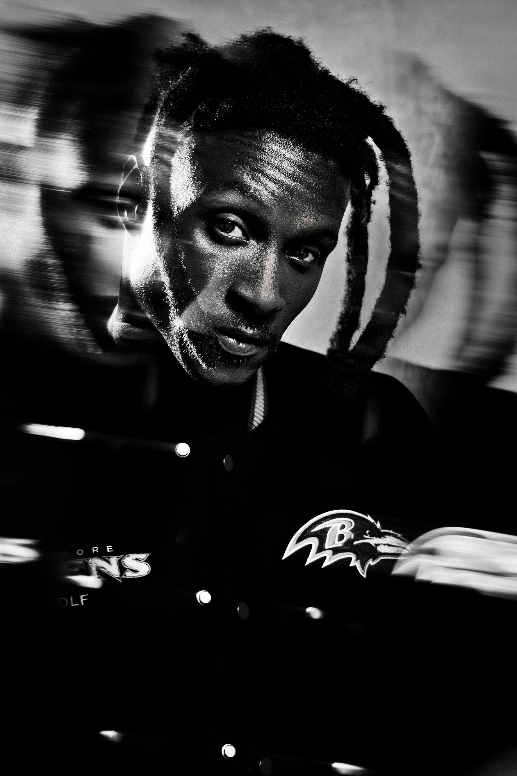 A black and white image of a woman with short curly hair, wearing a Baltimore Ravens jacket, looking directly at the camera with a serious expression.