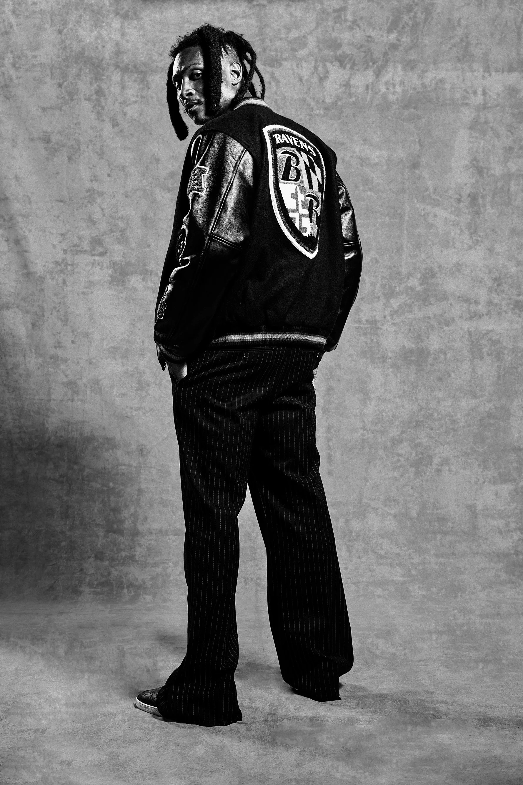 A person with dreadlocks wearing a black varsity jacket with a Baltimore Ravens logo on the back, black pinstripe pants, and black shoes, standing against a textured gray background.