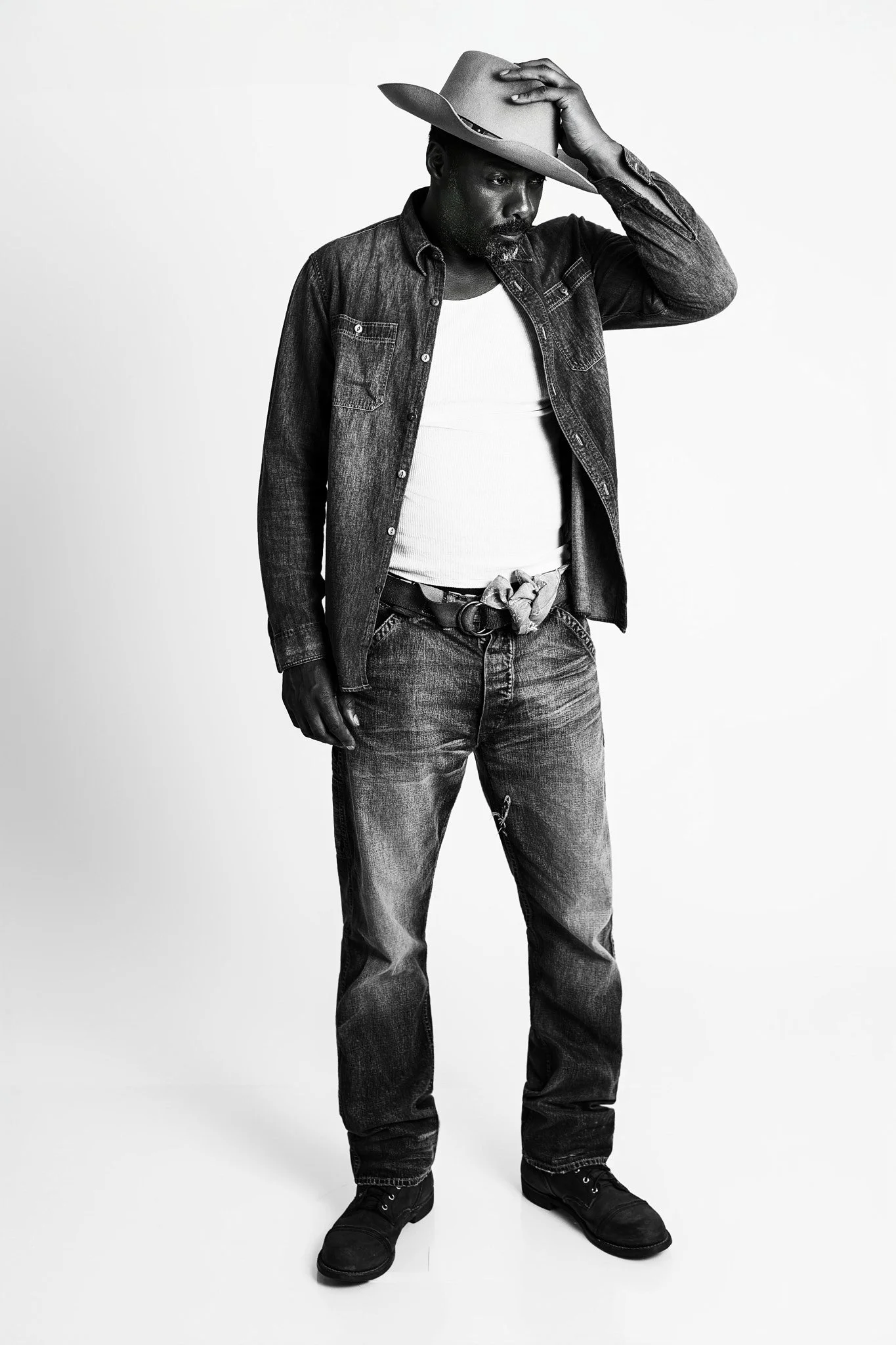 Black and white photo of a man wearing a cowboy hat, denim jacket, t-shirt, jeans, and boots, standing against a plain background and touching his hat with one hand.