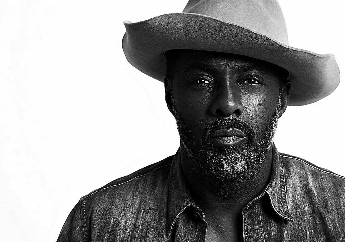Idris Elba wearing a wide-brimmed hat, denim shirt, and sporting a beard, in black and white portrait.