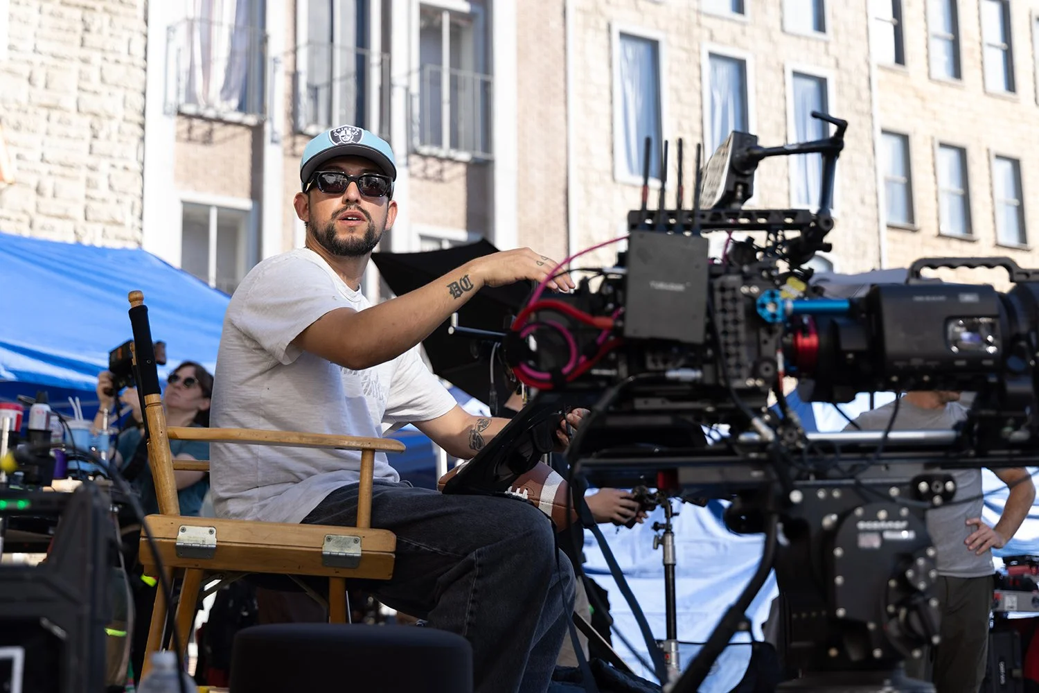 A man with a beard wearing sunglasses, a light-colored cap, and a white T-shirt sits on a wooden chair operating a professional film or video camera outdoors, with a city building in the background and other crew members around.