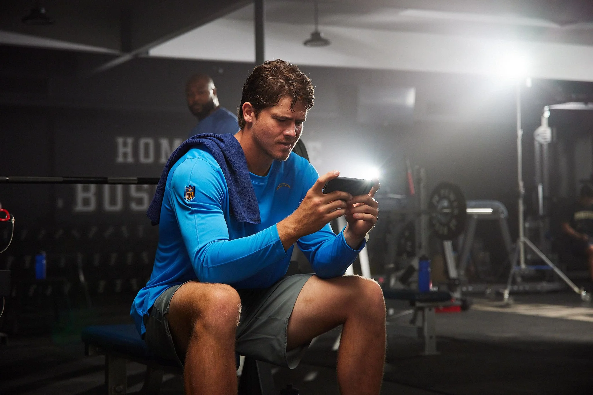 A young man in athletic clothes sitting on a bench in a gym, looking at his phone with a towel draped over his shoulders, with gym equipment blurred in the background.