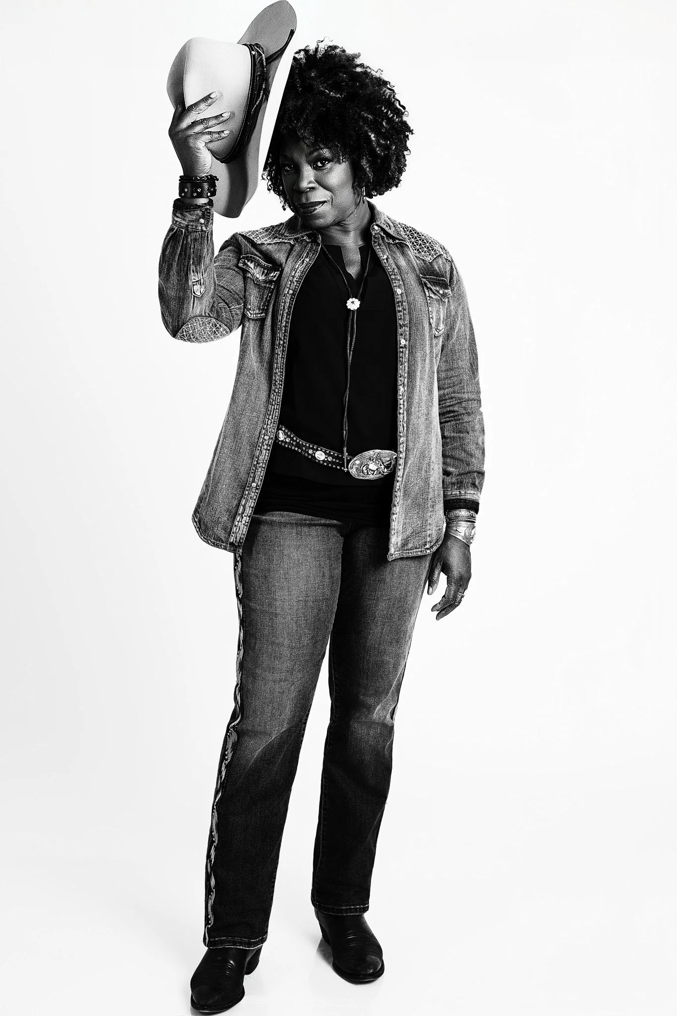 A woman with curly hair wearing jeans, a black top, and a denim jacket, holding a hat in her hand, standing against a plain white background.