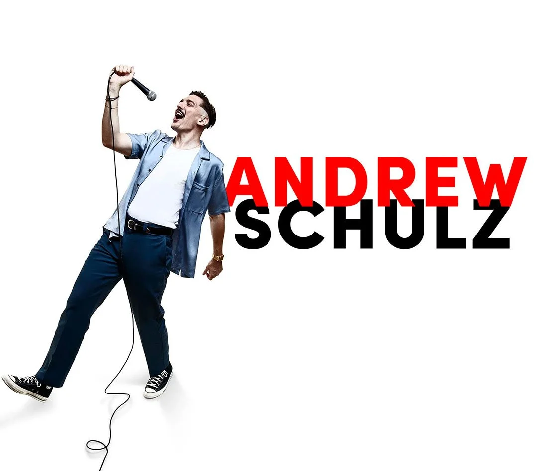 A man in a blue shirt and jeans singing into a microphone with the text "Andrew Schulz" in bold red and black letters.