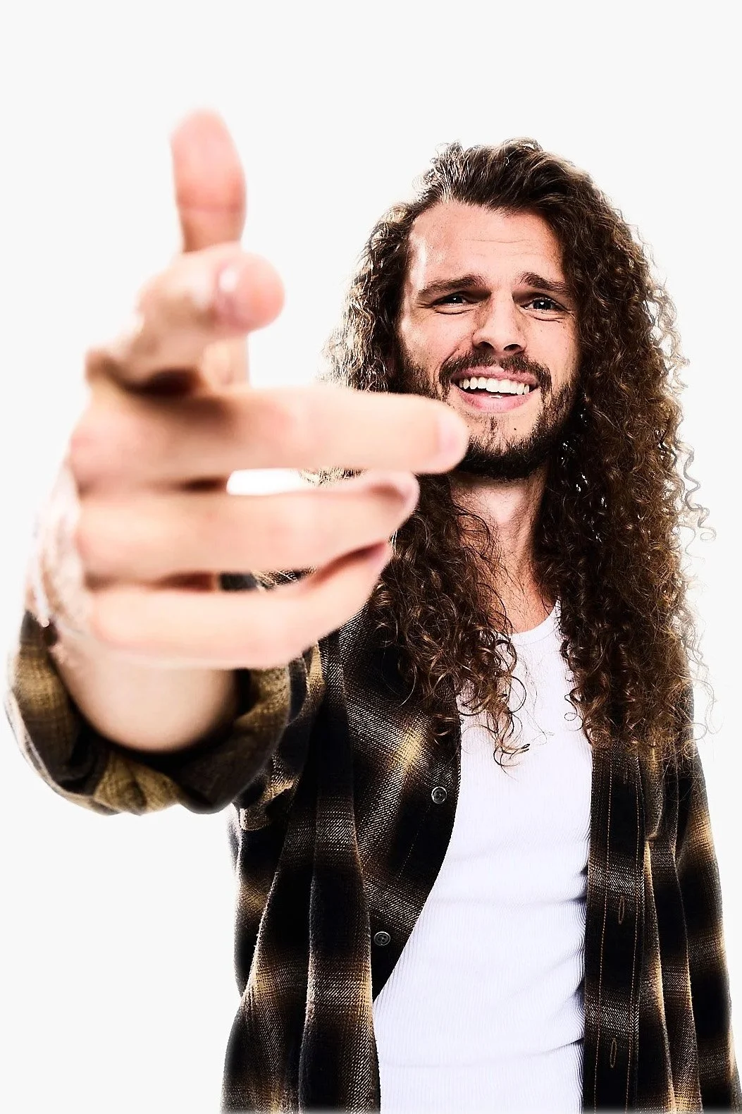 A man with long curly hair and a beard is smiling and pointing towards the camera, wearing a plaid shirt over a white t-shirt.