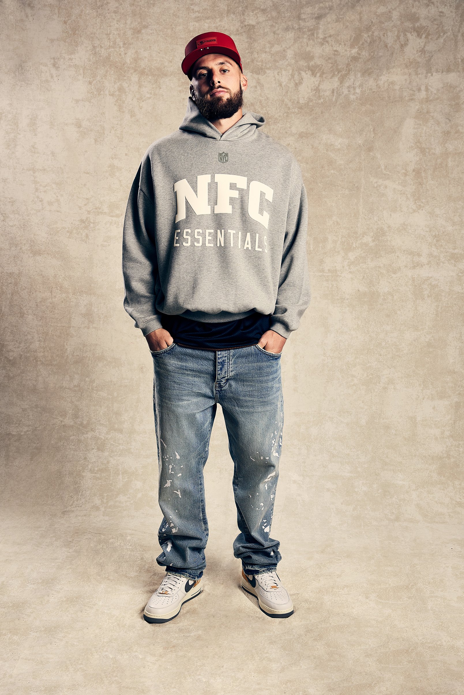 A young man wearing a red cap, gray hooded sweatshirt with 'NFC ESSENTIALS' on it, baggy jeans with paint splatters, and white Nike sneakers standing against a plain beige background.