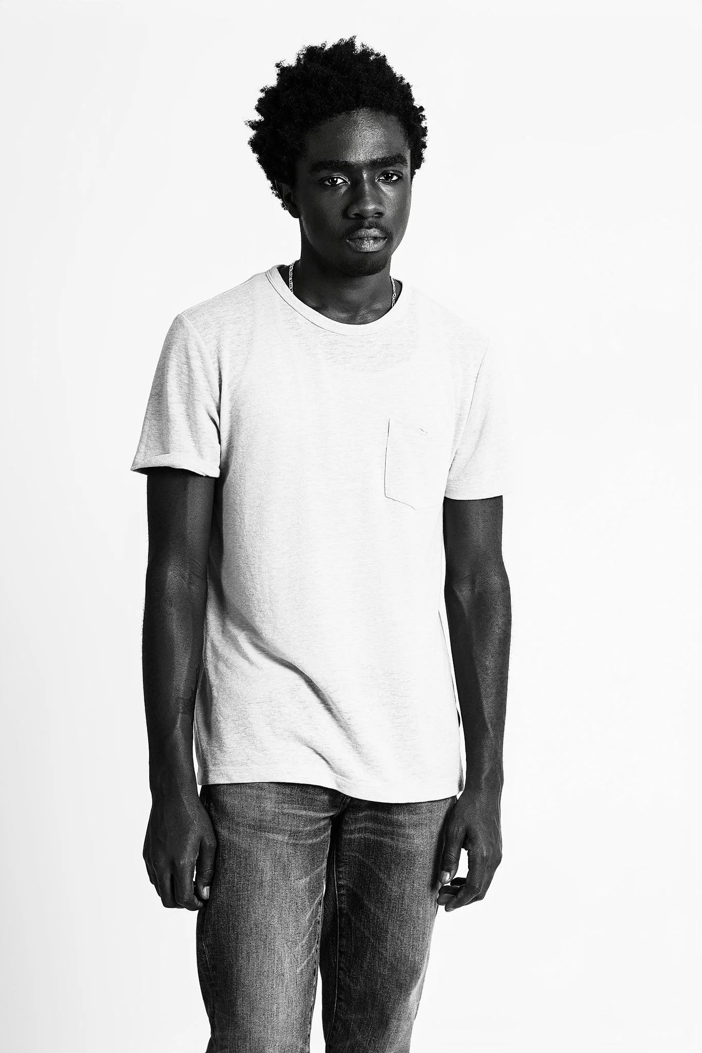 Black and white photo of a young man with curly hair wearing a light-colored t-shirt and jeans, standing against a plain background.