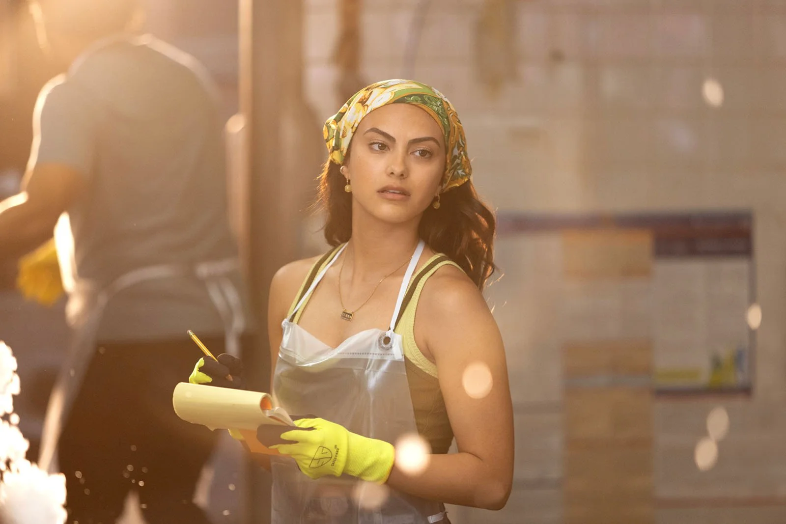 A young woman with a colorful headscarf, wearing a yellow tank top, an apron, yellow gloves, and jewelry, holding a notepad and pen in a bakery or kitchen environment, with a blurred background and warm sunlight.