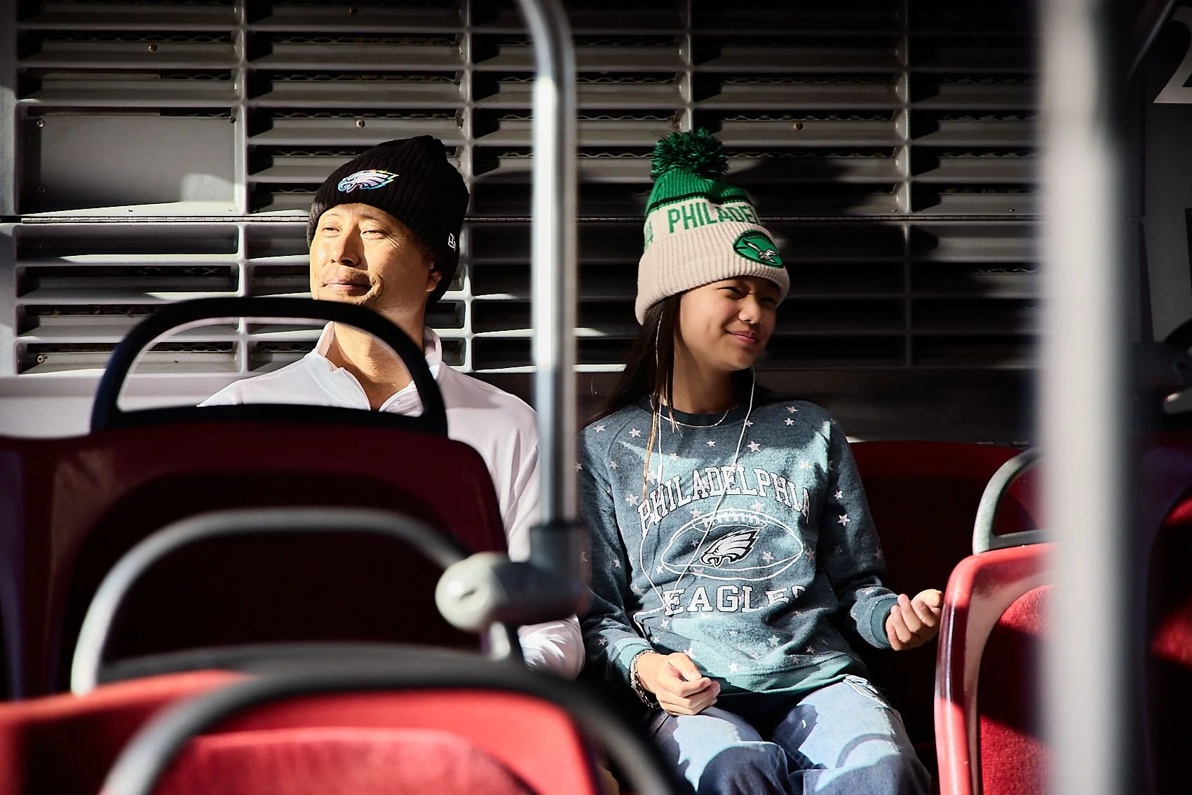 Two fans sitting on stadium seats, one man and one woman, both wearing Philadelphia Eagles merchandise, with sunlight on their faces.