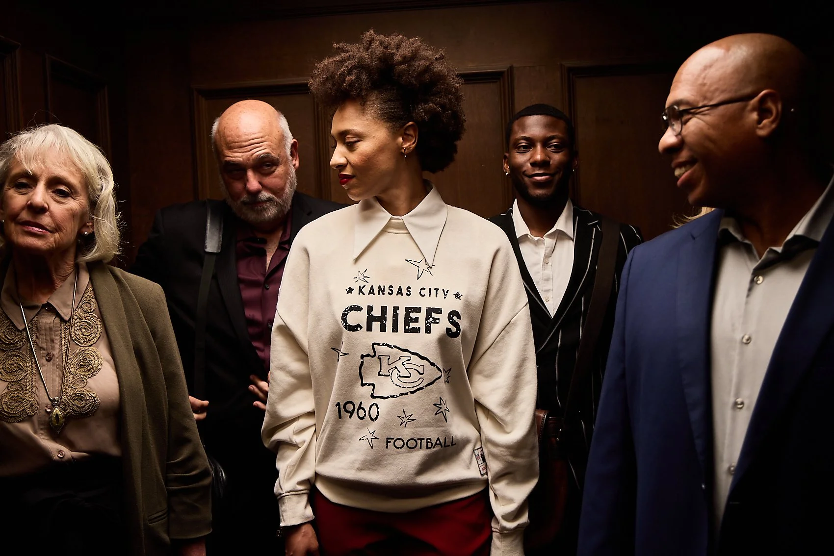 Group of five diverse adults, three women and two men, standing together in a wooden-paneled room, with one woman in the center wearing a vintage Kansas City Chiefs sweatshirt.