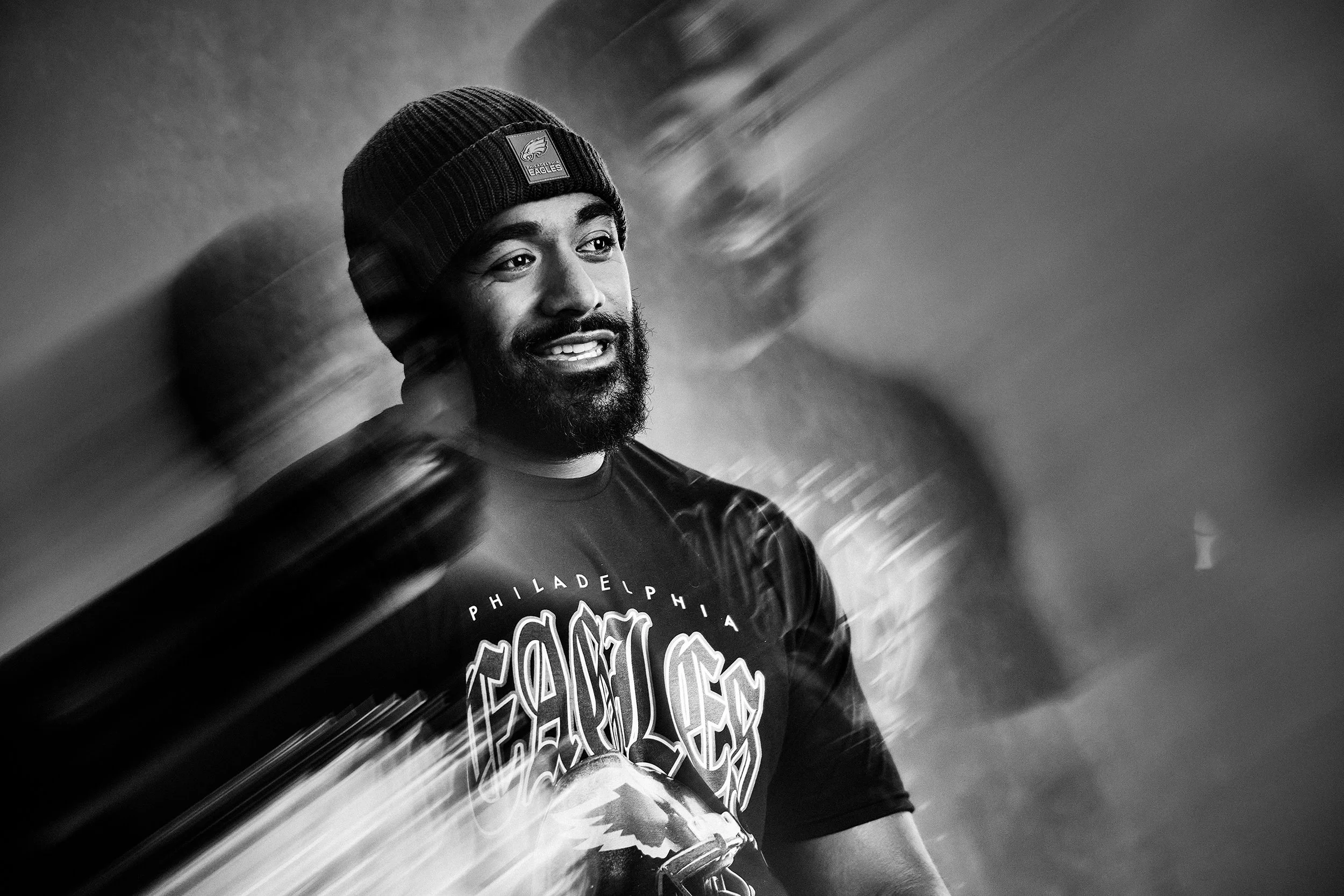 A smiling man with a beard and mustache wearing a Philadelphia Eagles beanie and a T-shirt with a graphic design, standing in front of a blurred background.