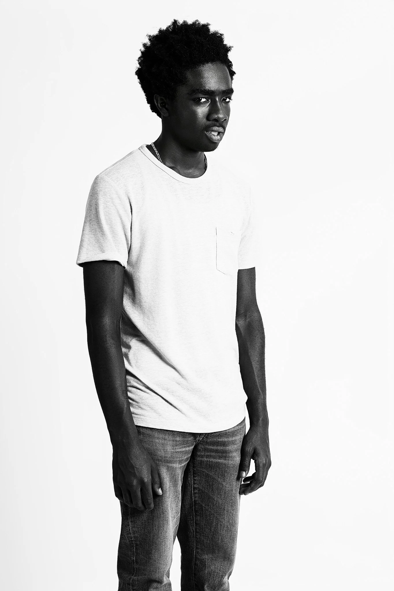 Black and white photo of a young man with curly hair, wearing a light t-shirt and jeans, standing against a plain background.