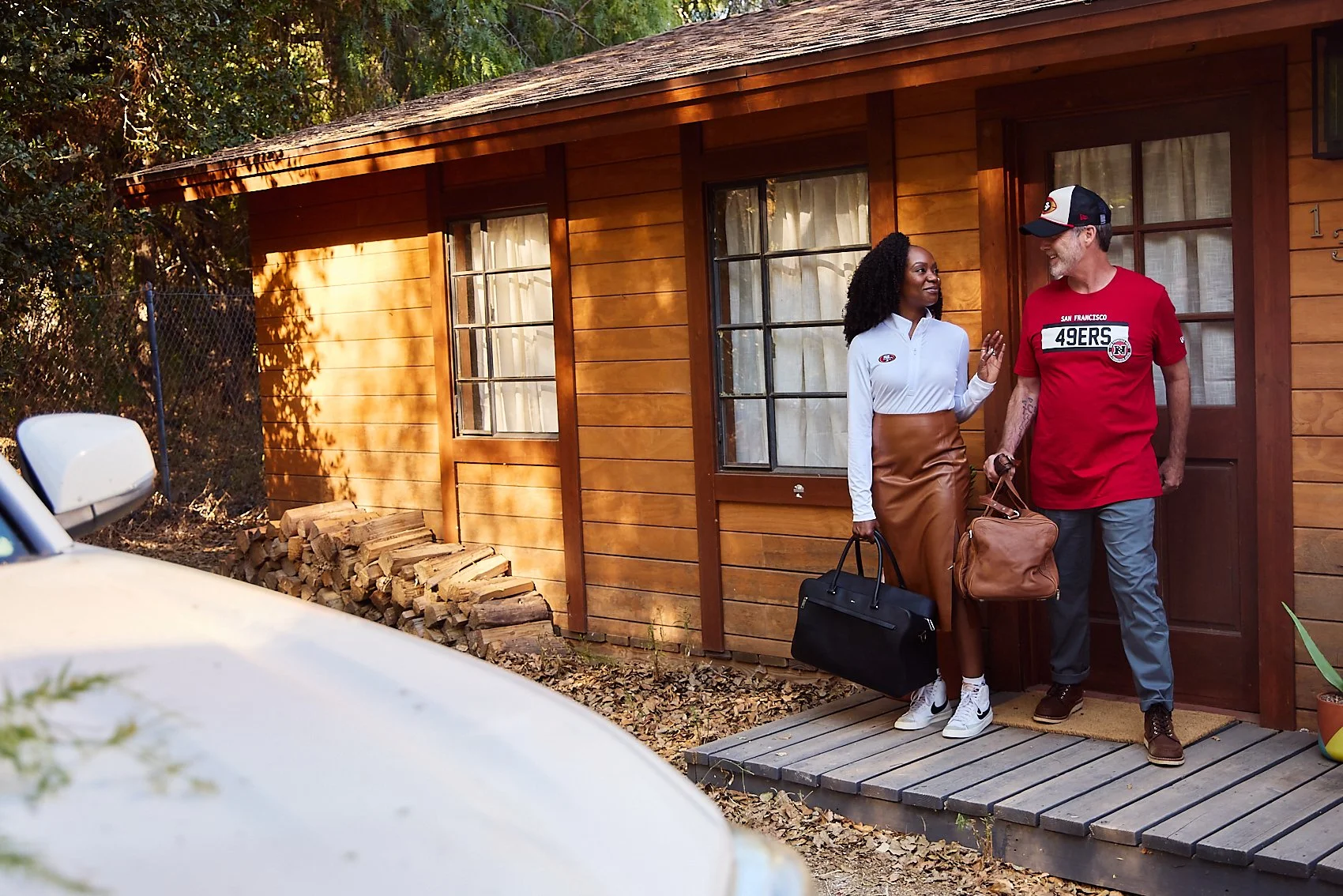 A man and woman talking on the porch of a wooden house, each carrying a bag, with a car visible in the foreground.