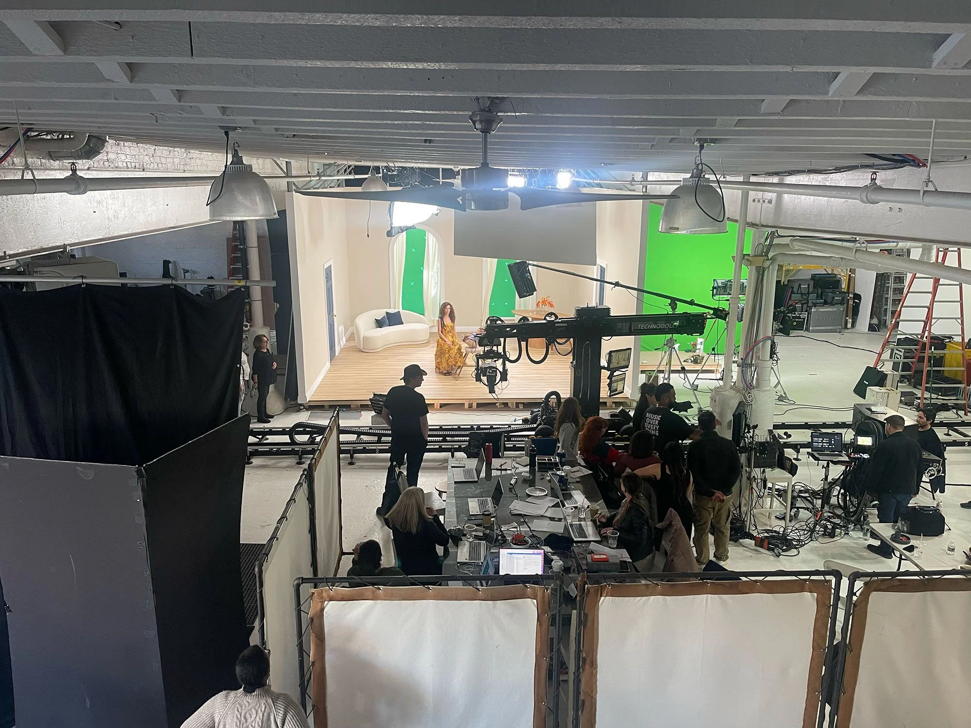 Inside a professional film or TV studio with a set featuring a woman seated on a stage with a white couch and green and white background panels. Crew members and equipment, including cameras, lights, and monitors, are visible, and a large black curtain is on the left side.