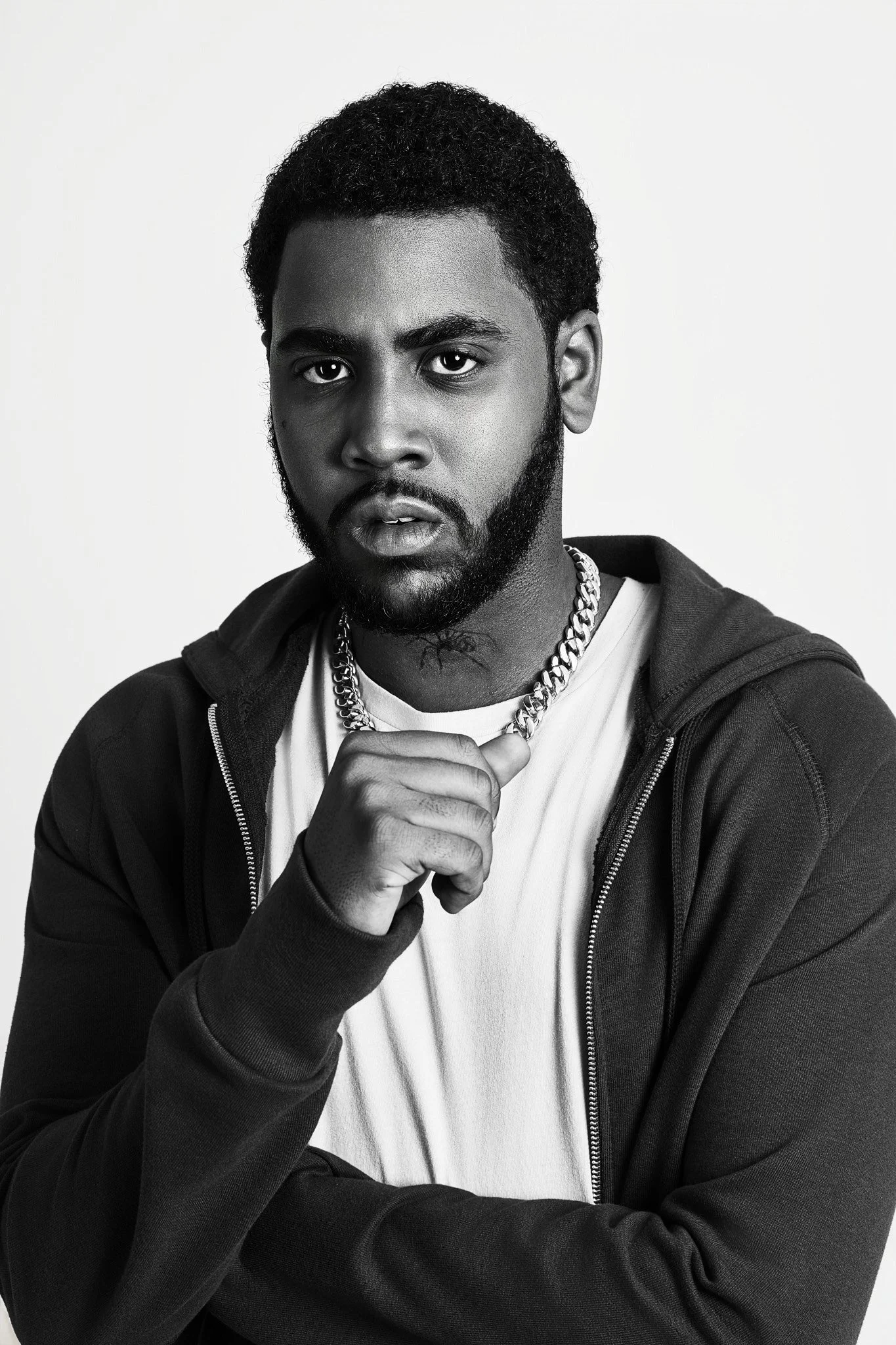Black and white portrait of a man with a beard and short curly hair, wearing a hoodie, white t-shirt, and a chain necklace, looking intense and serious.