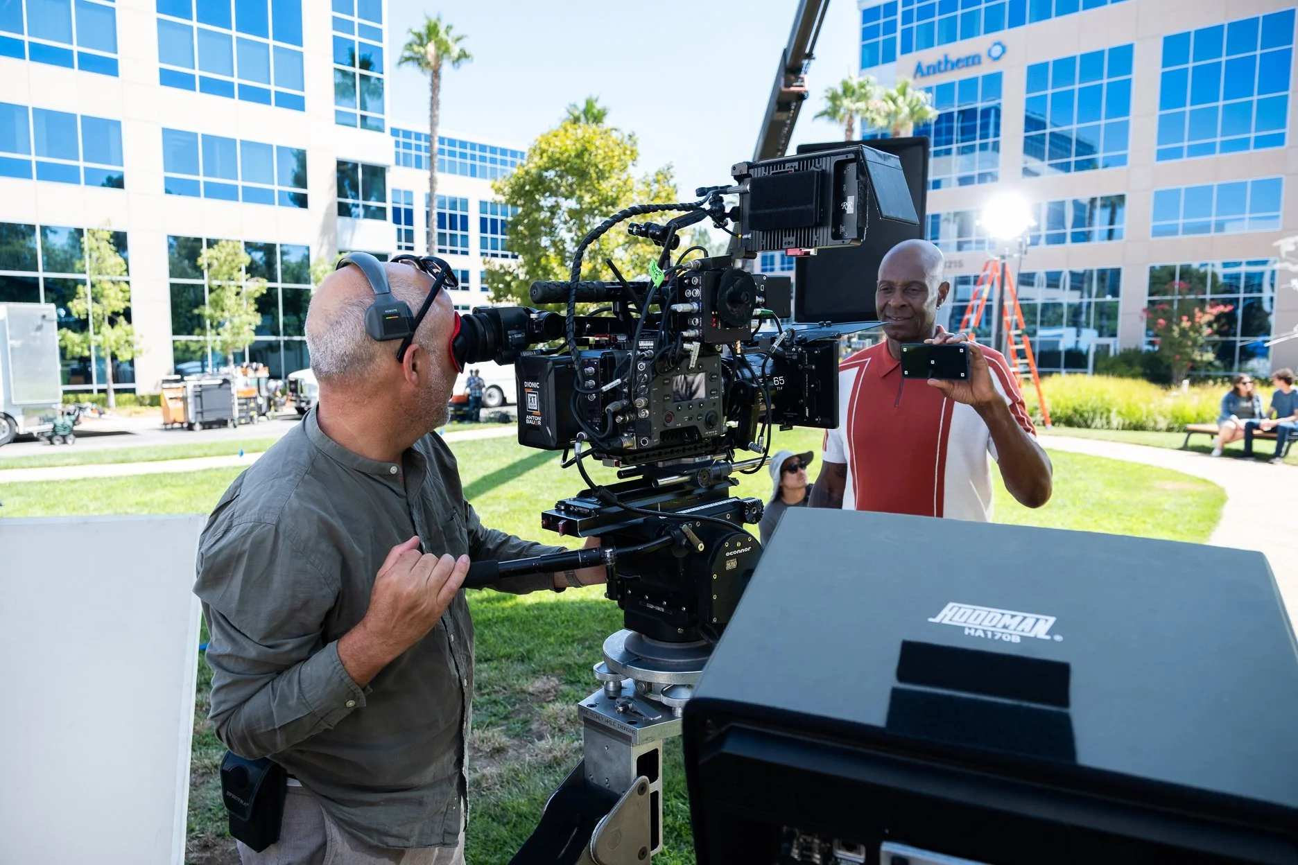 Filmmaker filming a man on a smartphone outdoors at a modern office building campus with a camera on a rig, sunlight, and green trees.