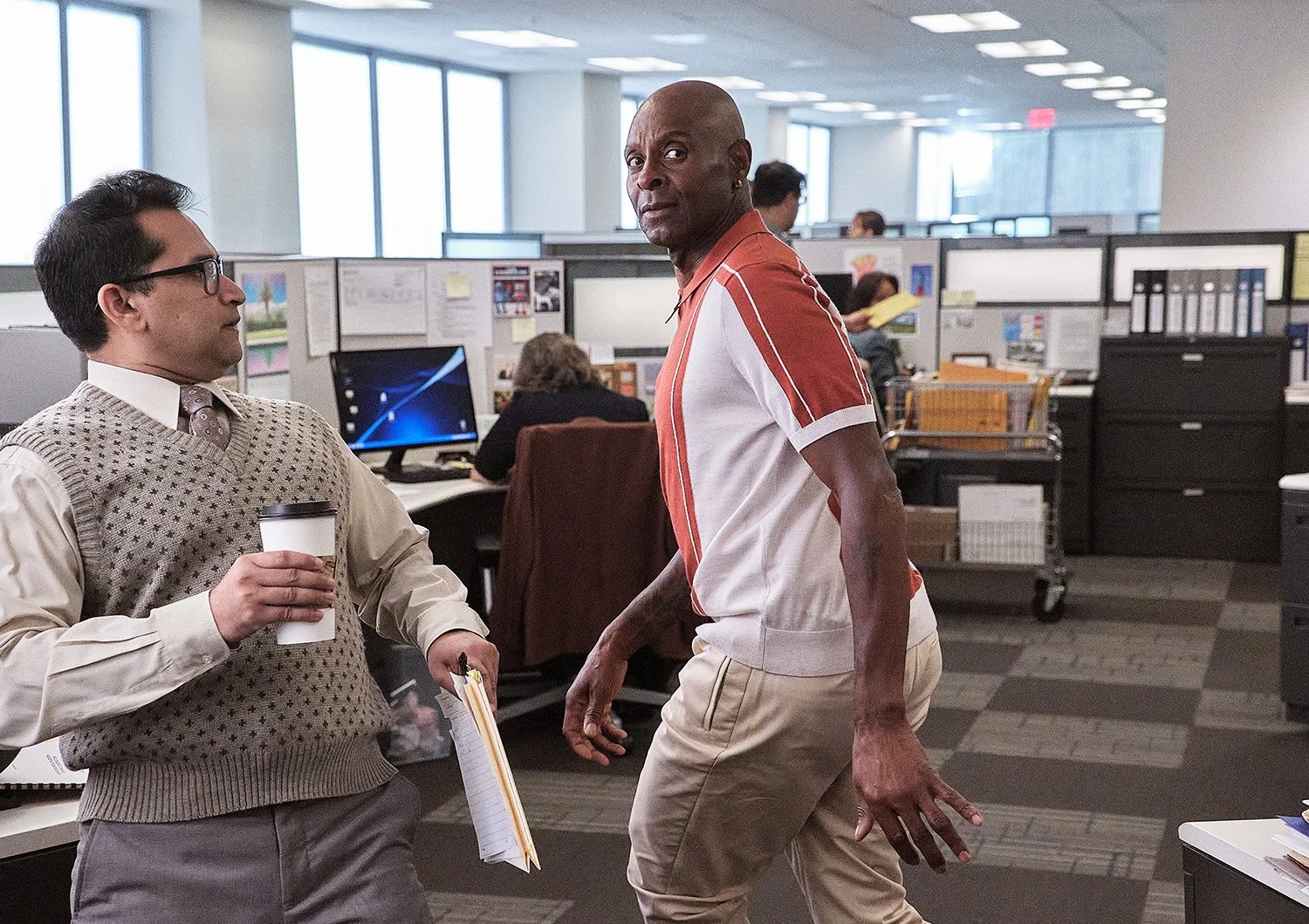 Two men in an office, one with glasses holding a coffee cup, and the other standing and looking at the camera. There are office cubicles and a woman working at a computer in the background.
