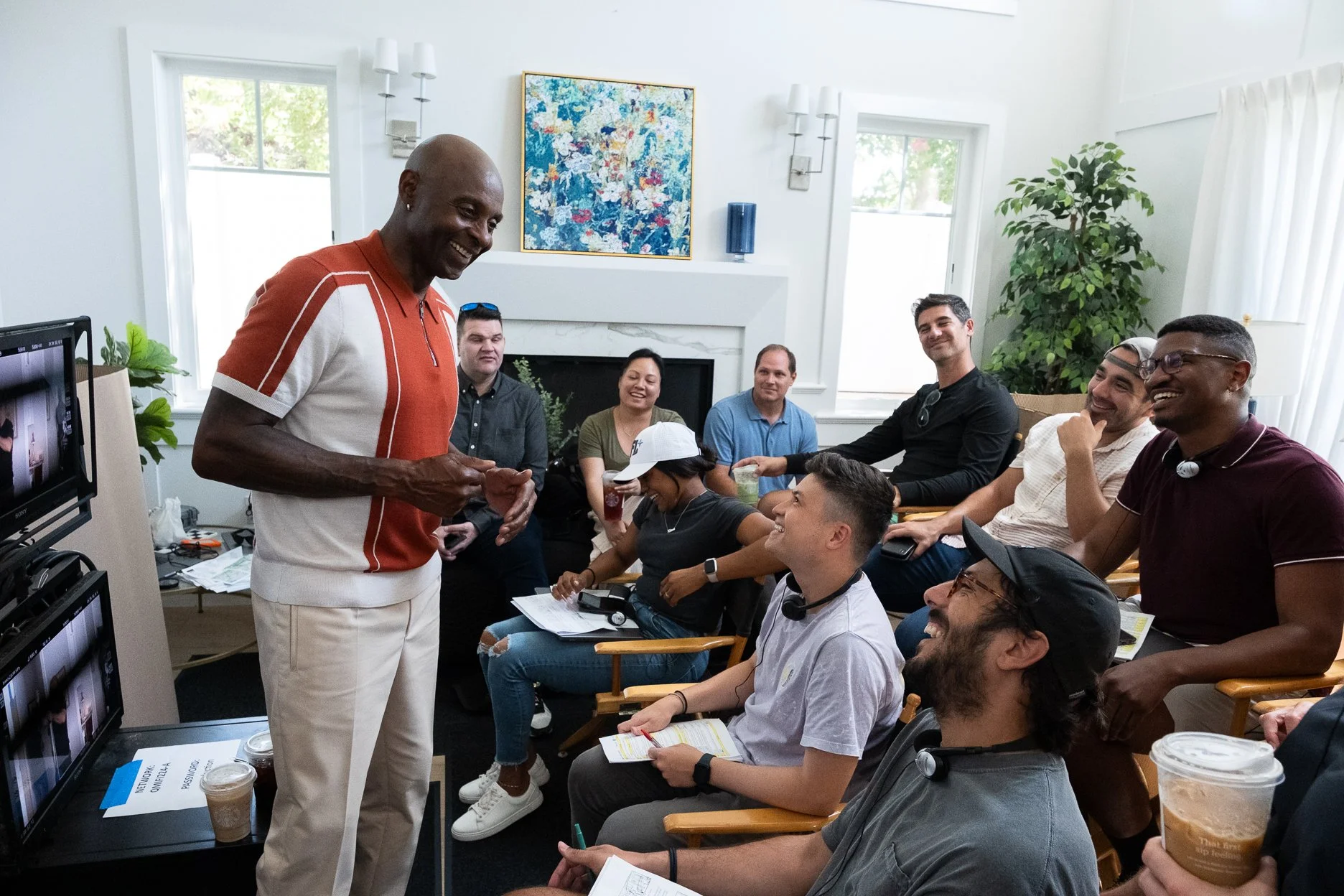 A man in a red and white shirt standing and smiling in front of a seated group of people in a bright living room, engaged in a discussion or presentation.