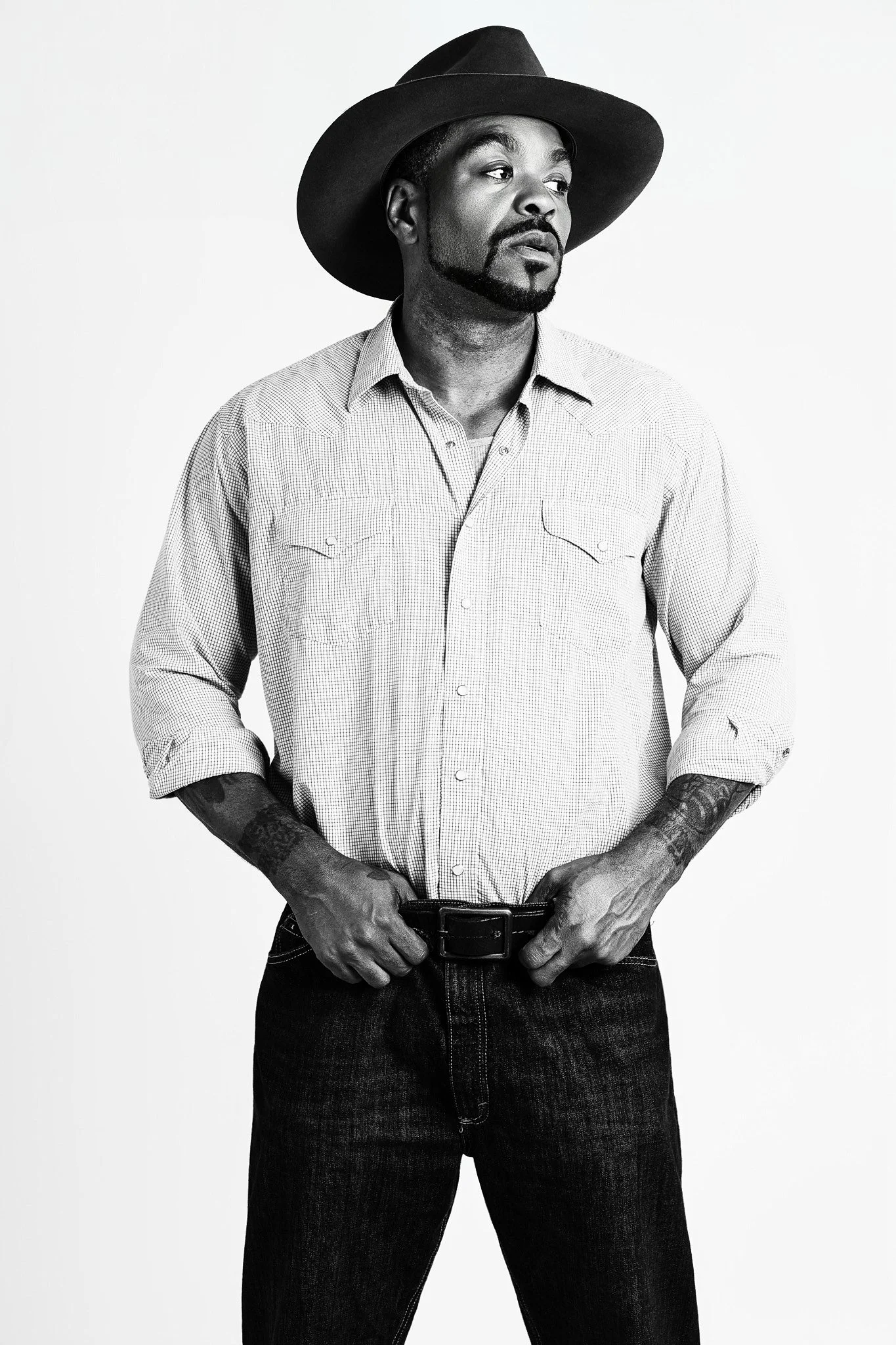 Black and white portrait of a man wearing a wide-brimmed hat, a button-up shirt, and jeans, standing with hands on his belt.