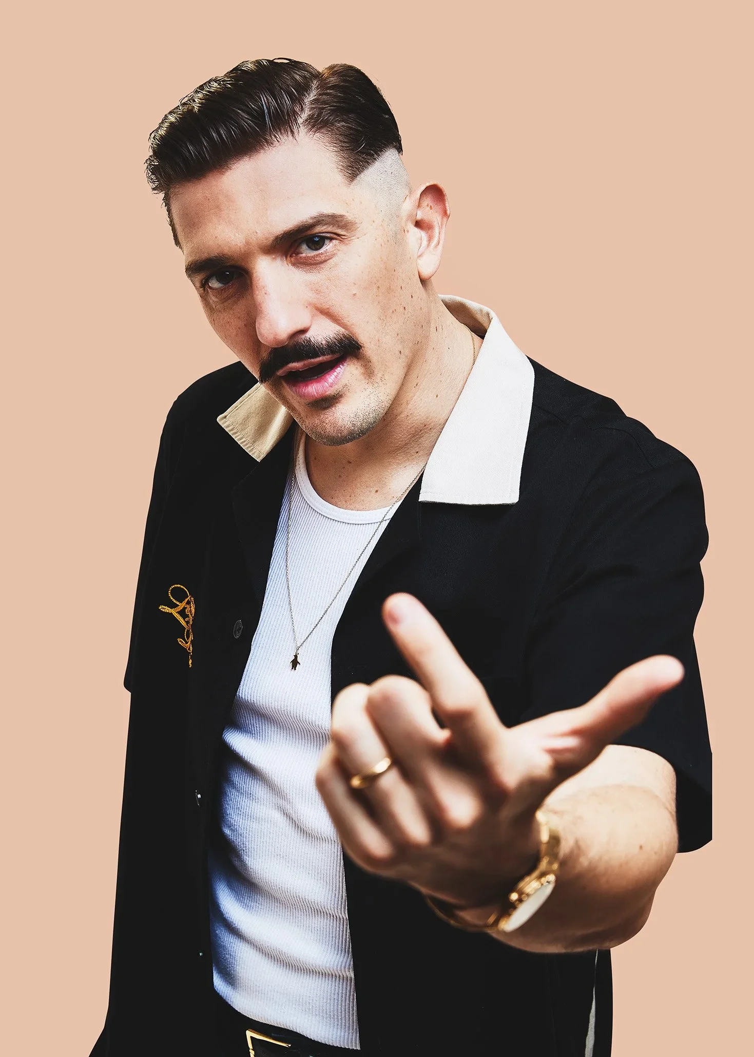 Man with dark hair and mustache, wearing a black shirt with white collar, pointing with his fingers, against a beige background.