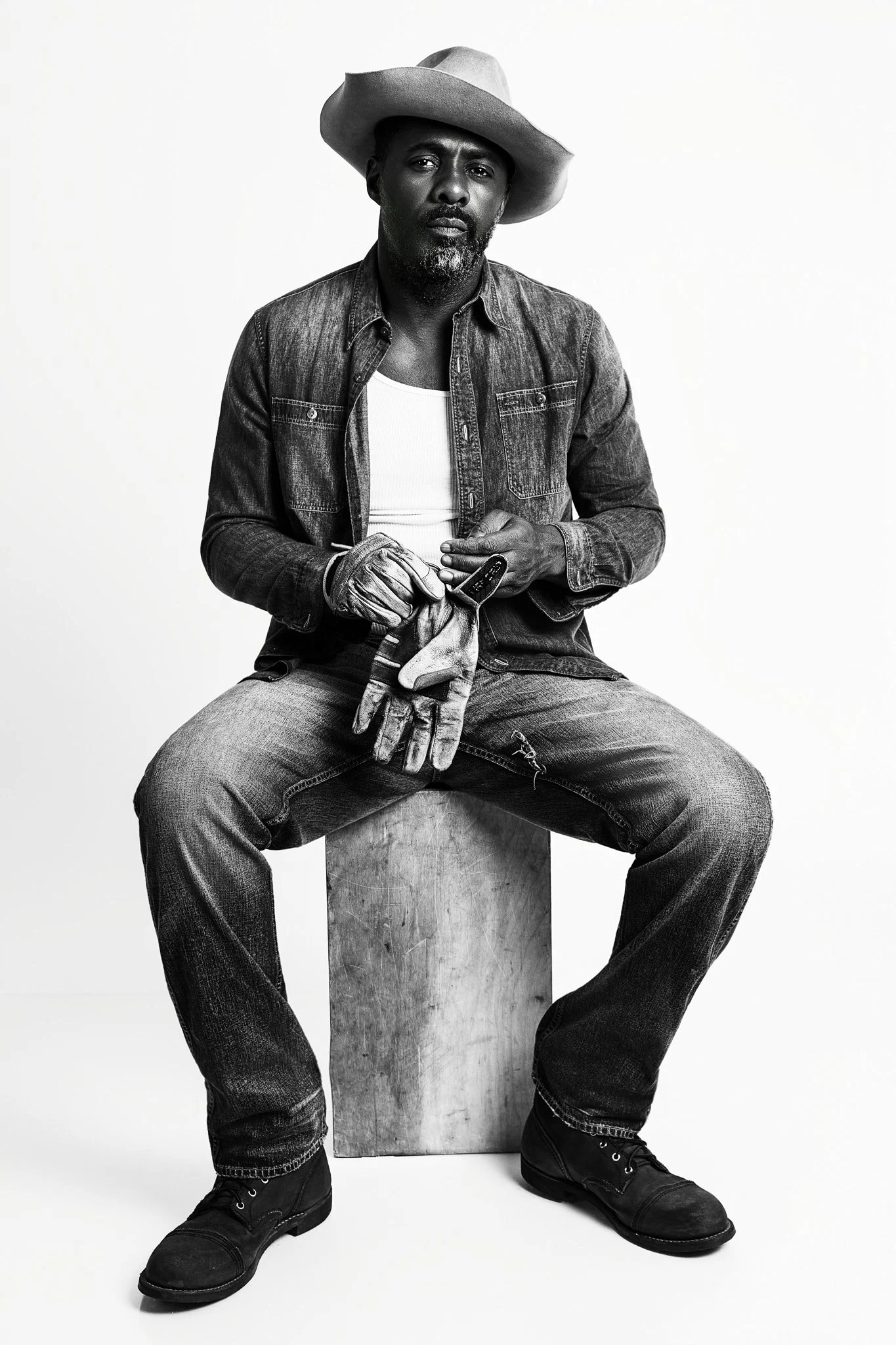 Black and white photo of a man sitting on a wooden block, wearing a cowboy hat, denim jacket, white tank top, jeans, and work boots, holding gloves and tools.