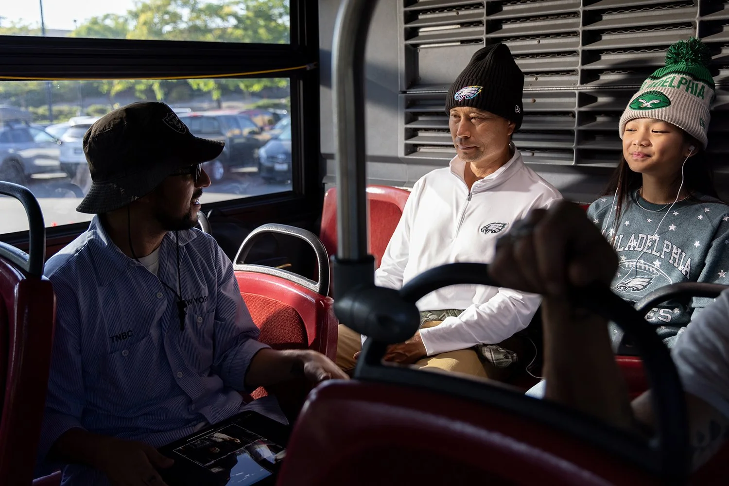 Three people sitting on a bus, with two men wearing Philadelphia Eagles hats and a girl wearing a gray Philadelphia sweatshirt and a green Eagles beanie, all listening to music or podcast.