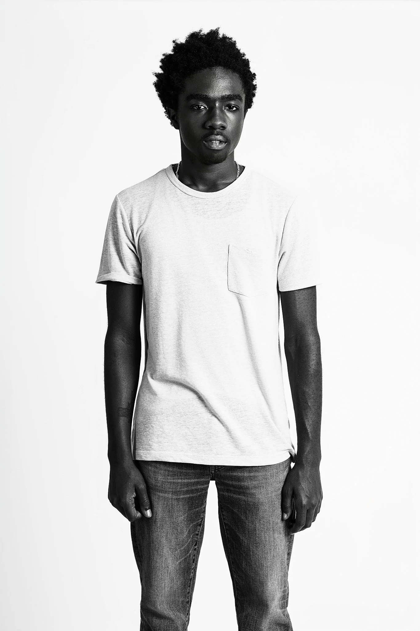 Black and white photo of a young man with curly hair, wearing a light-colored t-shirt and jeans, standing against a plain background.