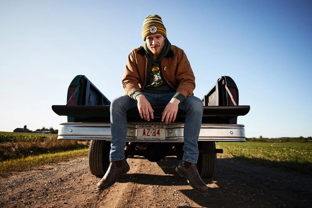 A man sitting on the edge of a pickup truck bed with a rural landscape in the background, wearing a beanie, jacket, and jeans.