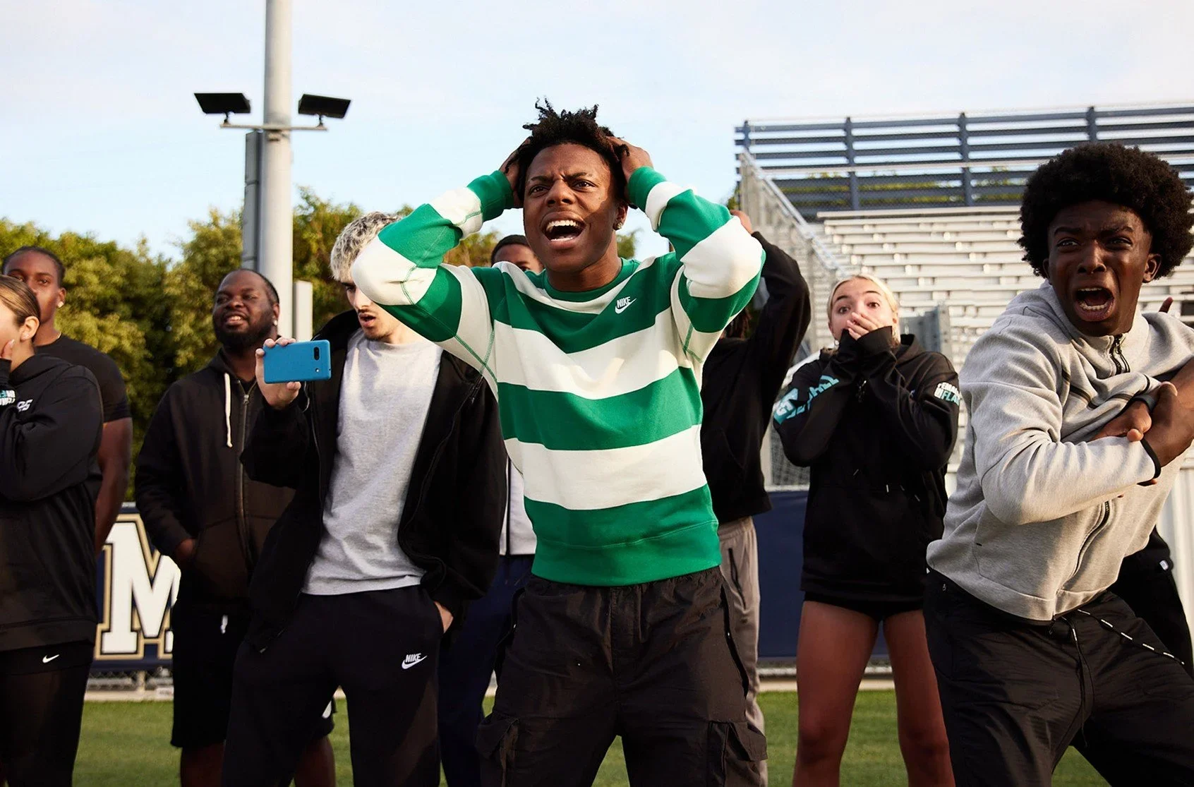Group of people outside on a field, displaying reactions of shock and concern, with one person in a green and white striped shirt holding his head.