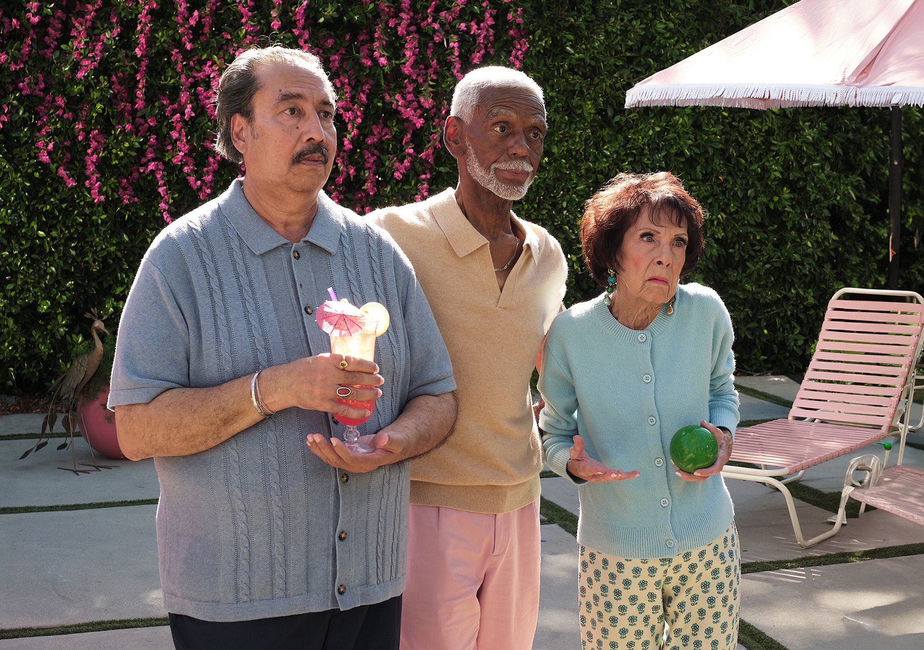 Three older adults standing outdoors, the man on the left holding a cocktail with a pink umbrella, the man in the middle with gray hair and beard, and a woman on the right holding a green ball, with pink flowers and greenery in the background, and pi