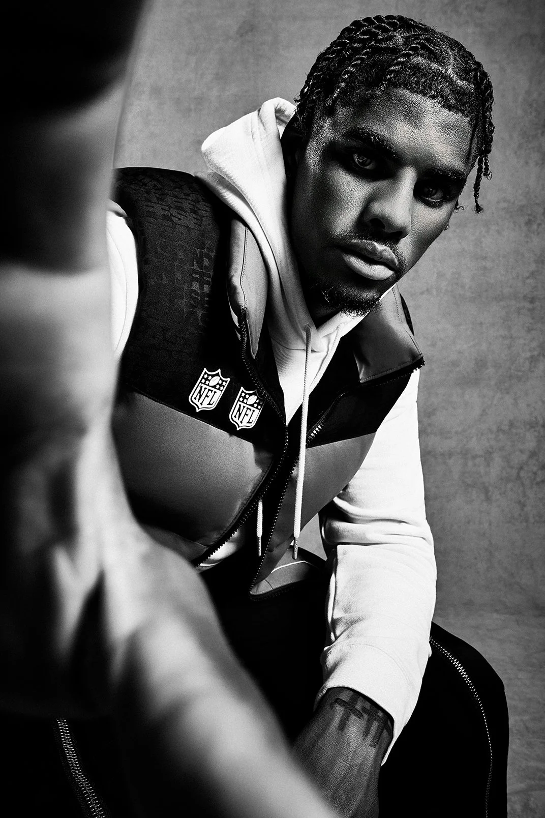 A black and white photograph of a man with braided hair, wearing a NFL vest and hoodie, looking directly at the camera against a plain background.