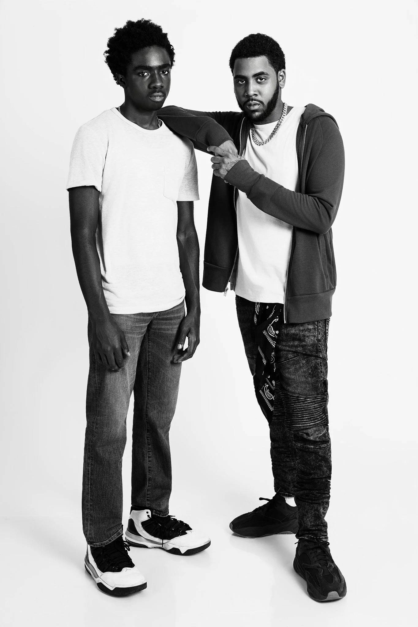 Two young men standing together in a studio, one with his arm around the other's shoulder, both dressed in casual clothing and sneakers, black and white photo.